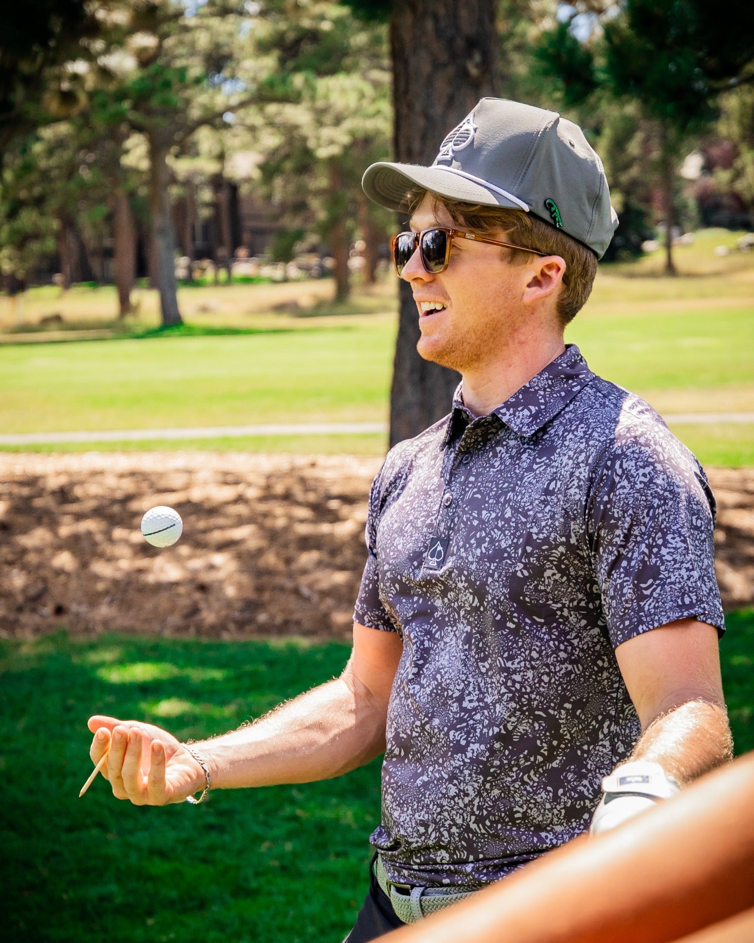 A golfer in a patterned shirt and cap juggles a golf ball on a sunny course, showcasing a playful moment.