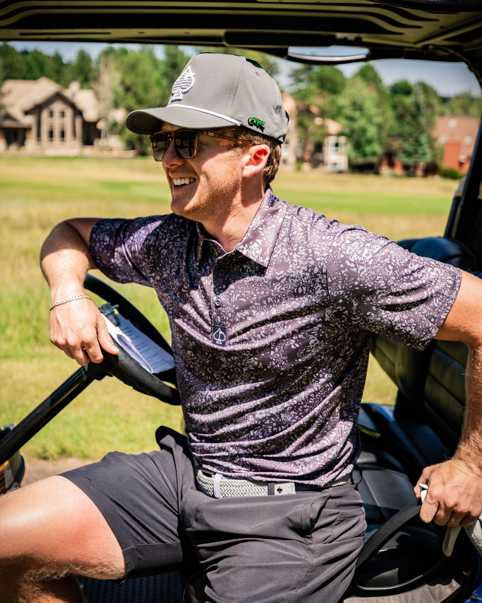 A golfer wearing a stylish purple and black patterned polo shirt and gray cap, smiling while seated in a golf cart on a sunny day.