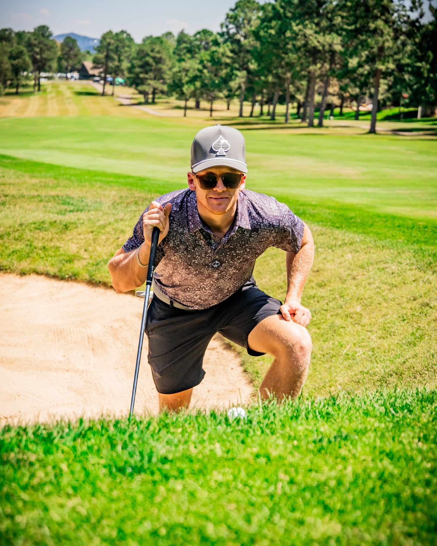 A golfer in a stylish shirt and cap climbs out of a sand bunker on a sunny golf course, showcasing bold golf fashion.