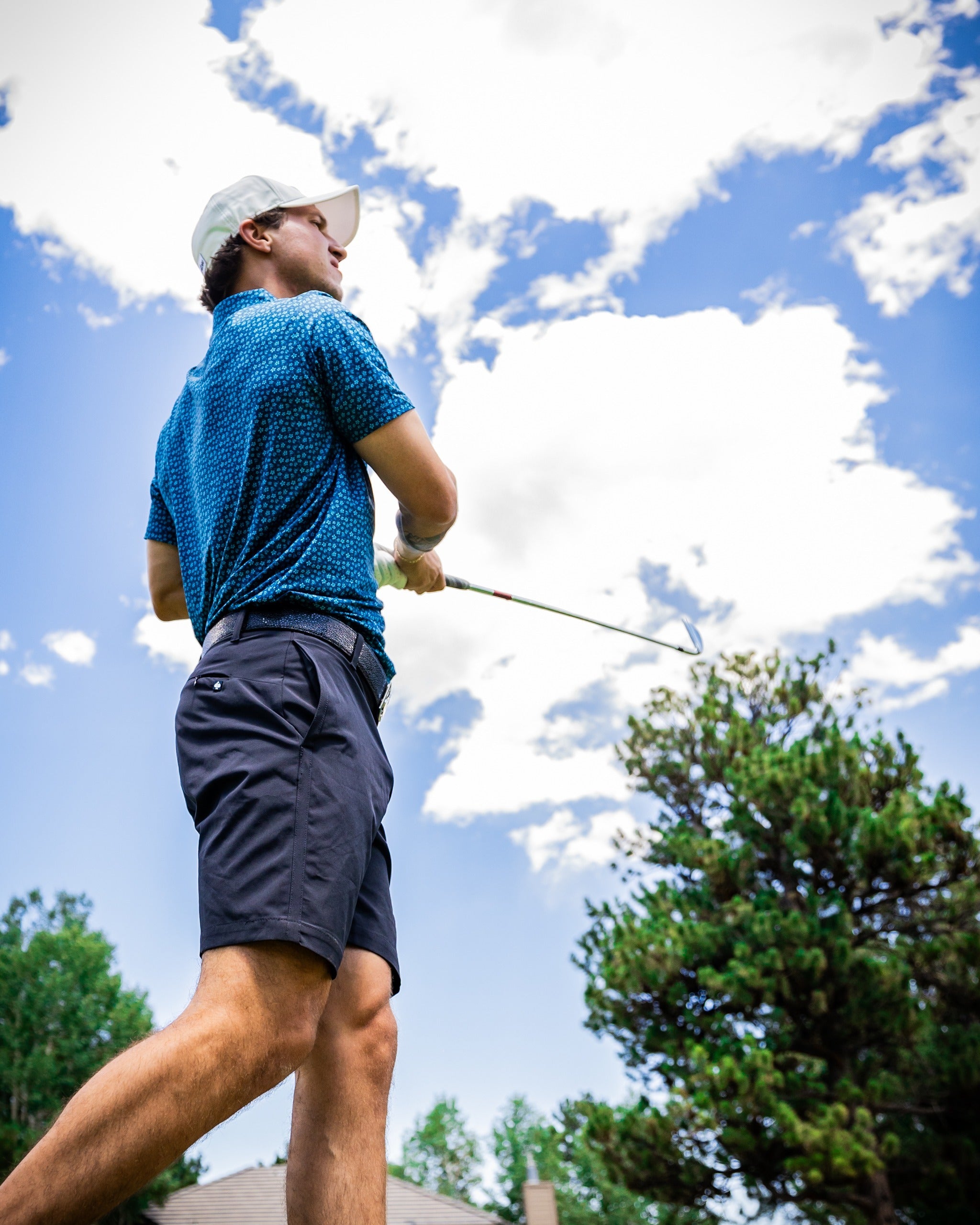 A golfer swings a club while wearing a blue micro floral polo shirt and shorts, set against a bright sky and trees.