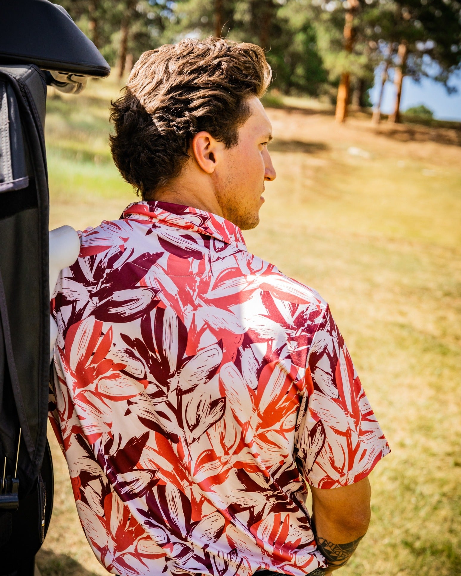 Man wearing a vibrant brushed floral golf shirt, leaning against a golf bag on a sunny course.