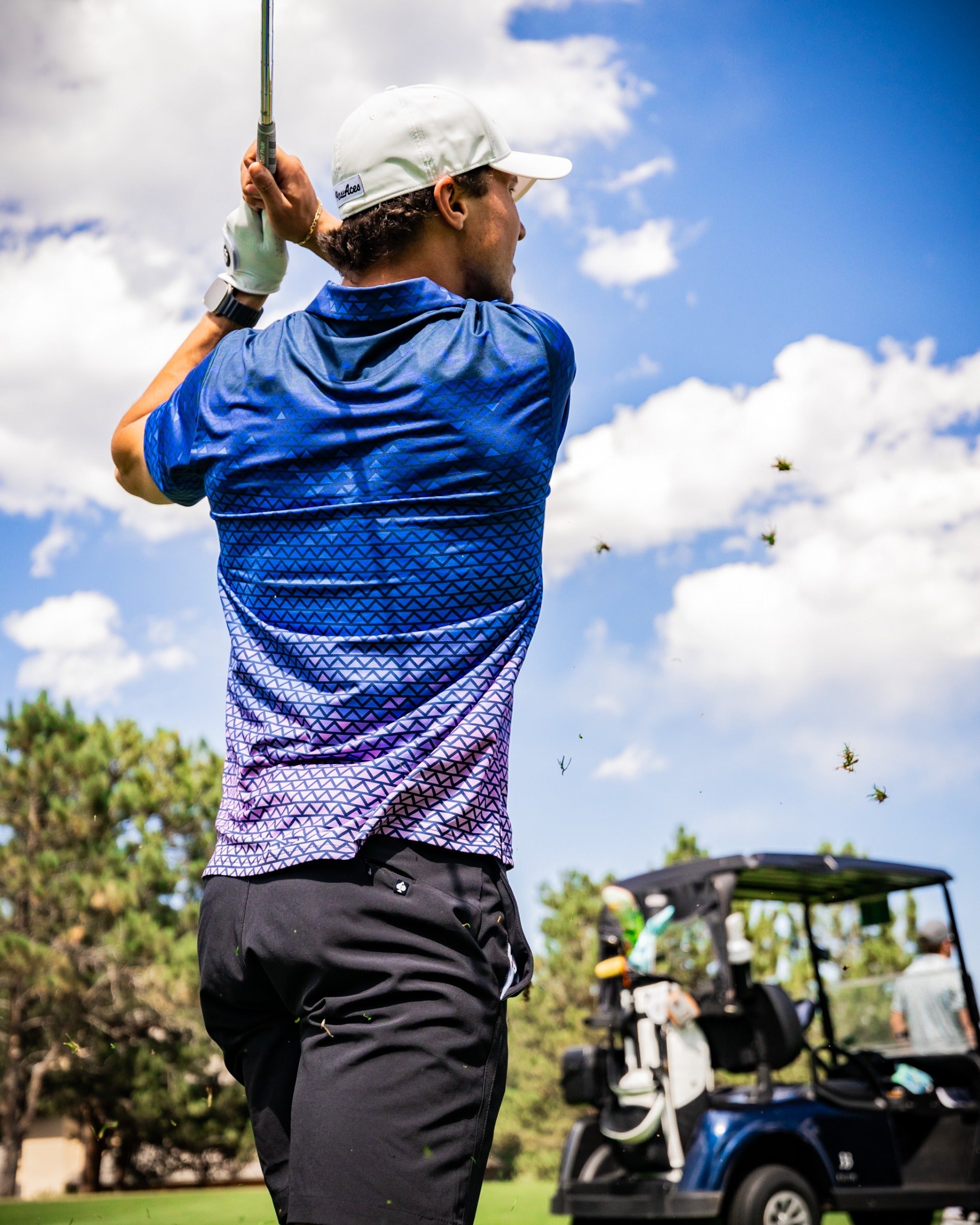 A golfer swings a club in a vibrant blue and purple patterned shirt, with a golf cart in the background under a sunny sky.