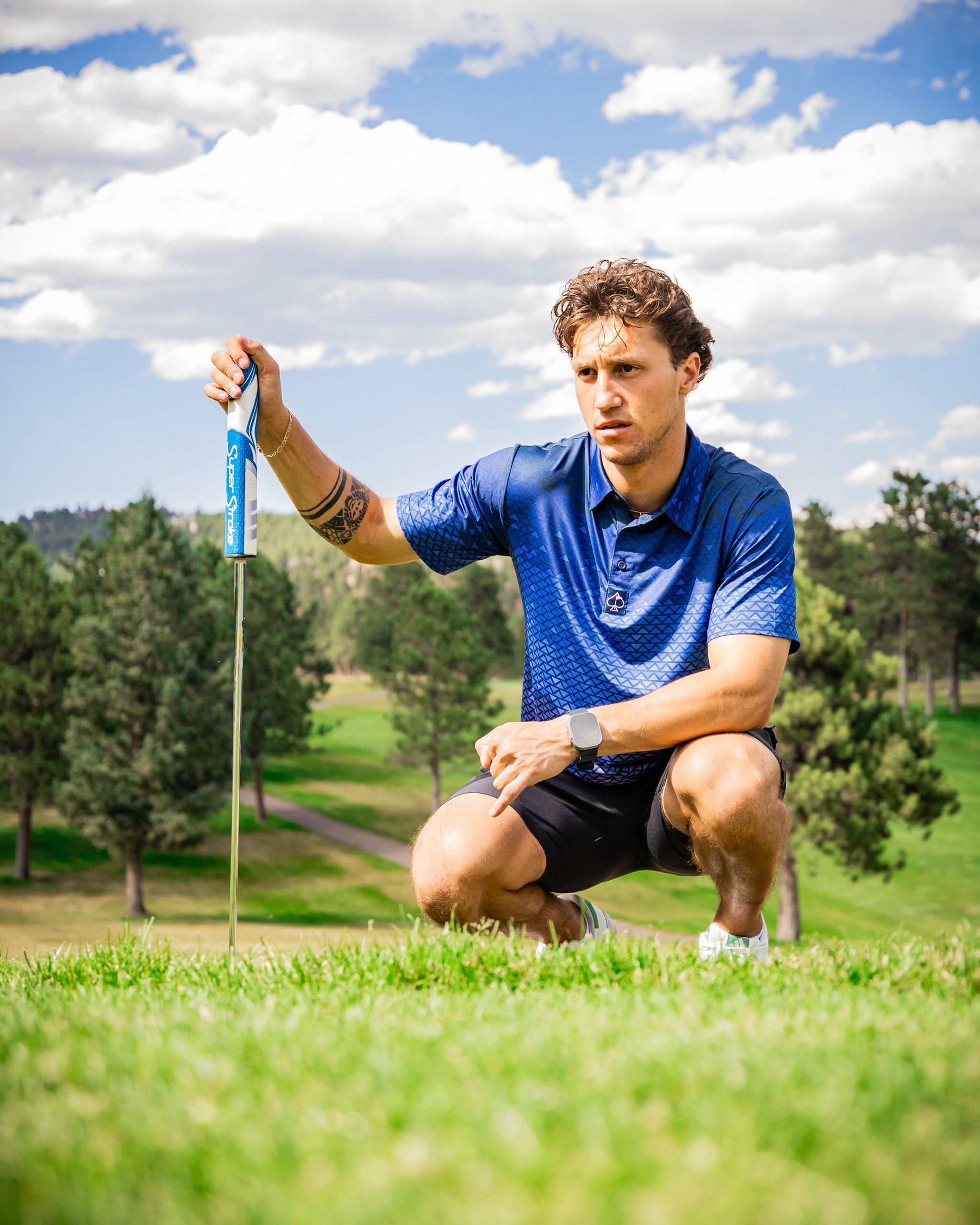 A golfer in a blue patterned polo shirt kneels on the green, focusing intently on his putt with a golf club in hand.