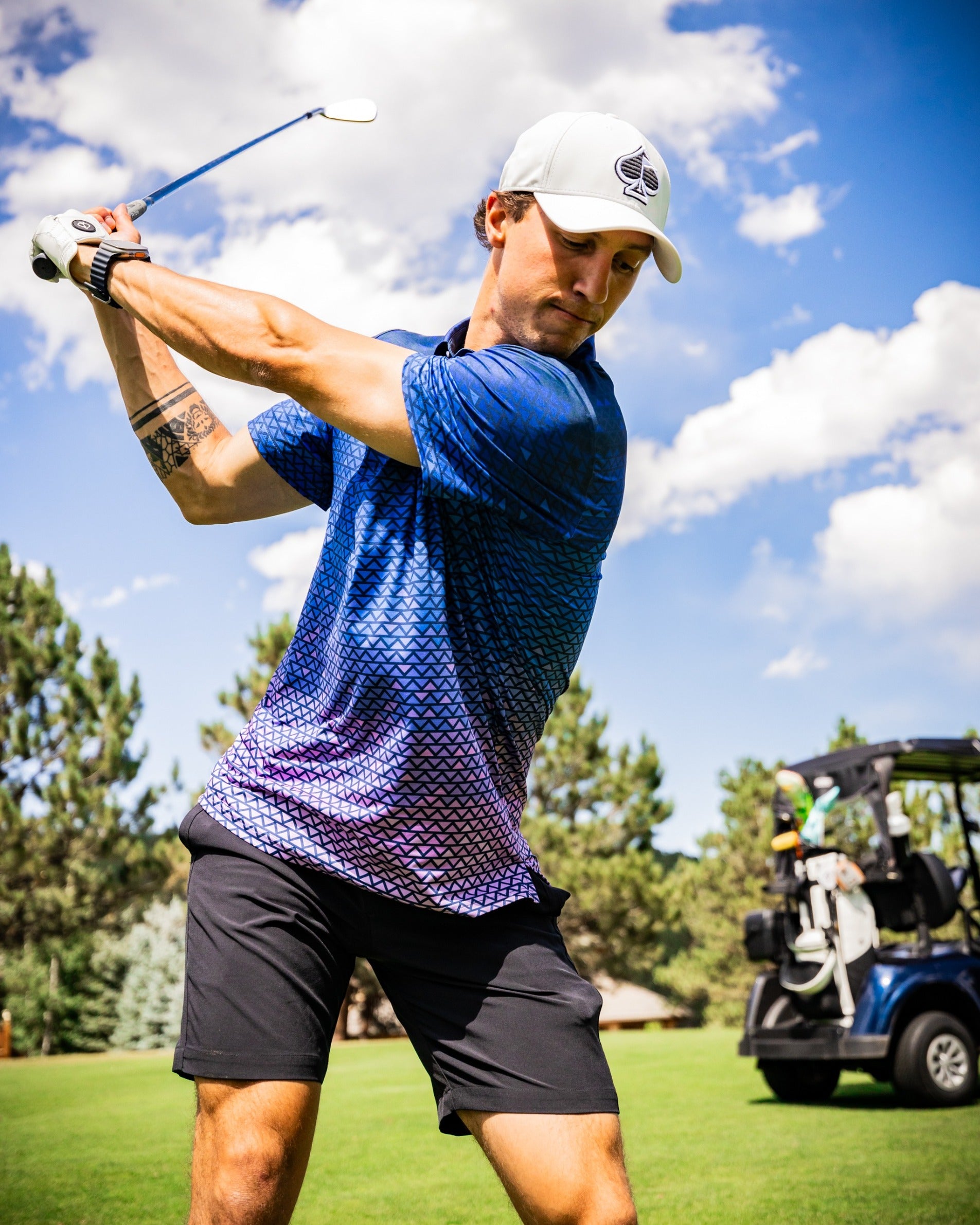 A golfer swings a club on the course, wearing a blue patterned polo shirt and black shorts, with a golf cart in the background.