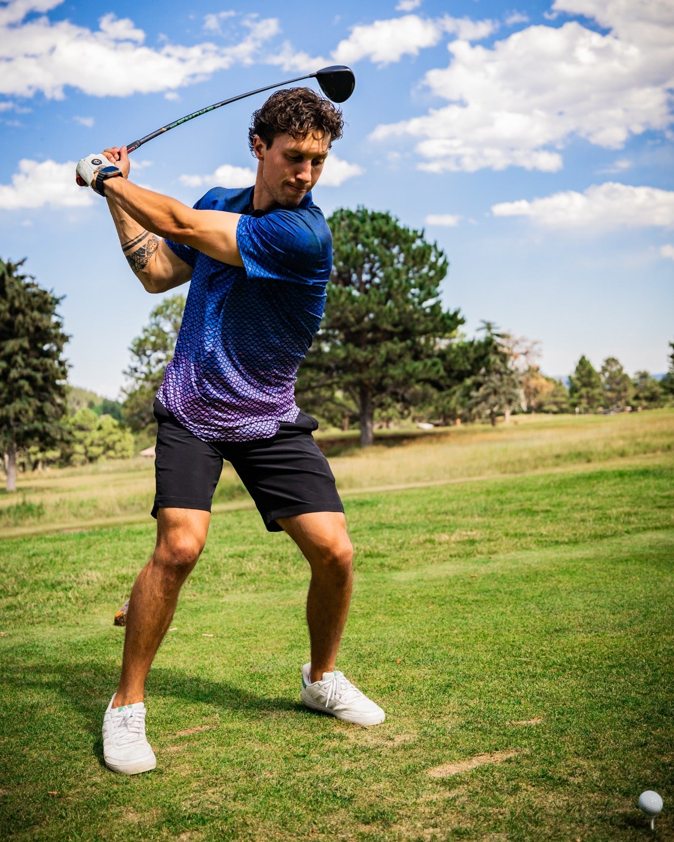 A golfer swings a club on a sunny day, wearing a blue and purple gradient shirt and black shorts on a green course.