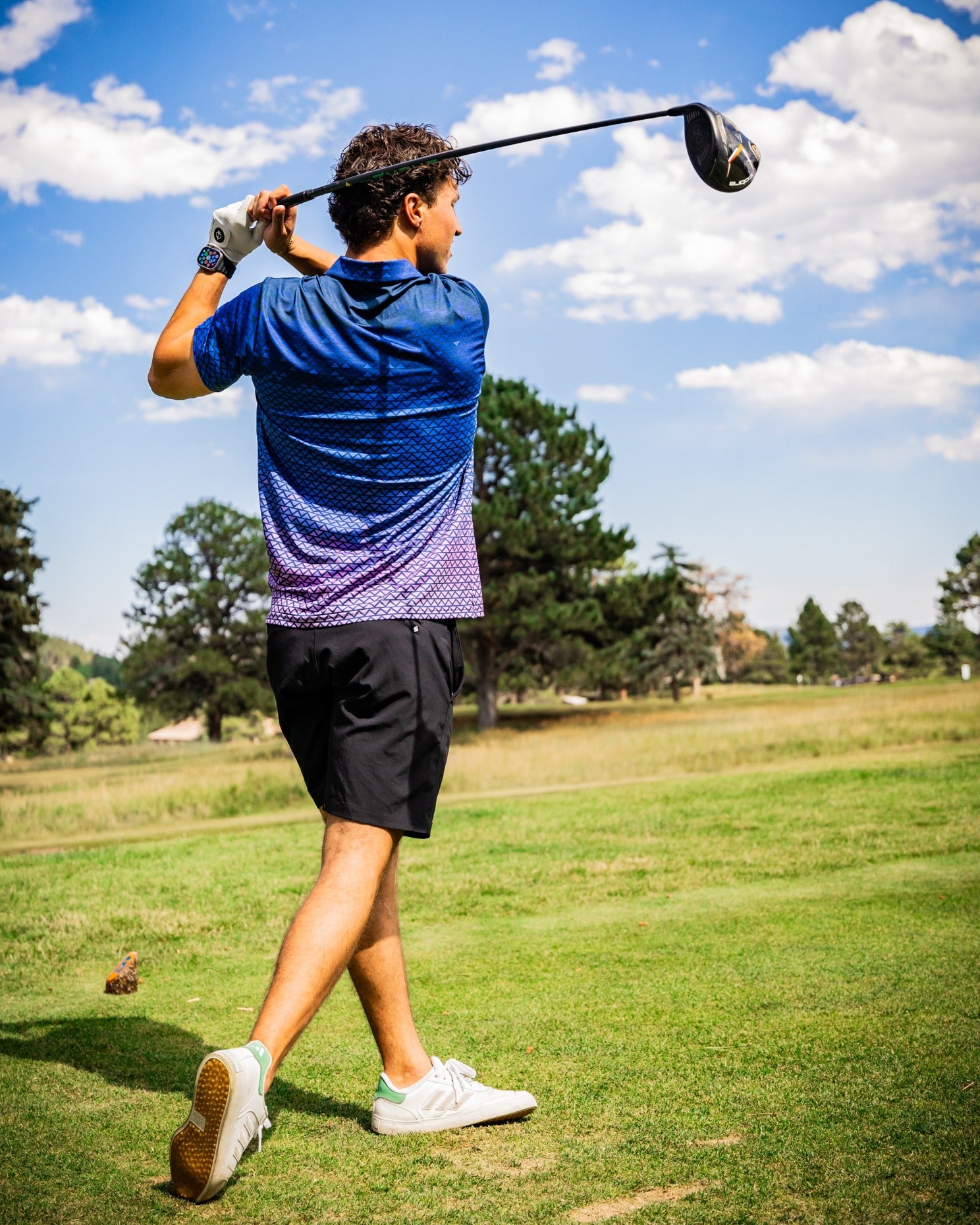 A golfer swings a driver on the course, wearing a blue and purple digital rainfall polo and black shorts.