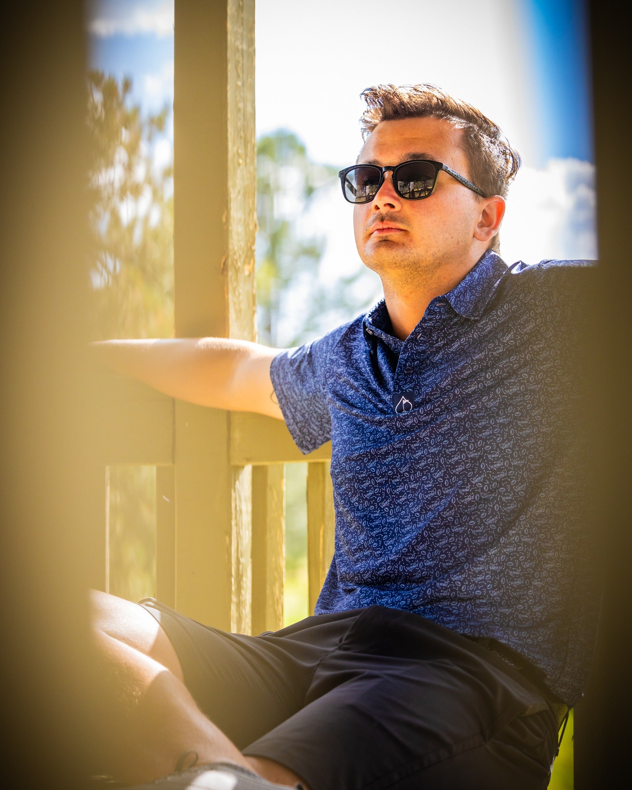 Man wearing a stylish blue patterned golf polo and sunglasses, relaxing outdoors in a sunny setting.