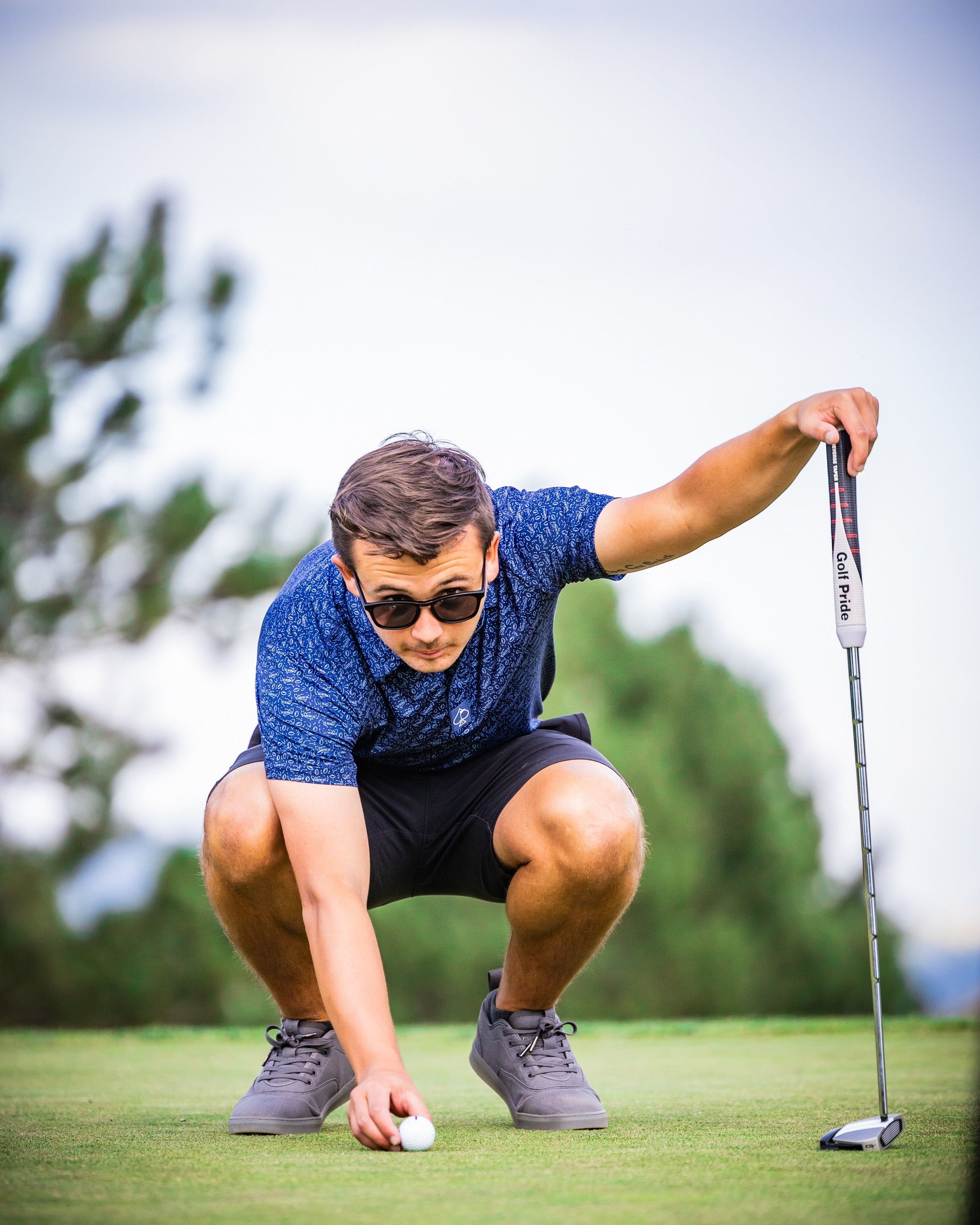 A golfer in a blue patterned shirt and sunglasses crouches on the green, lining up a putt with a putter.