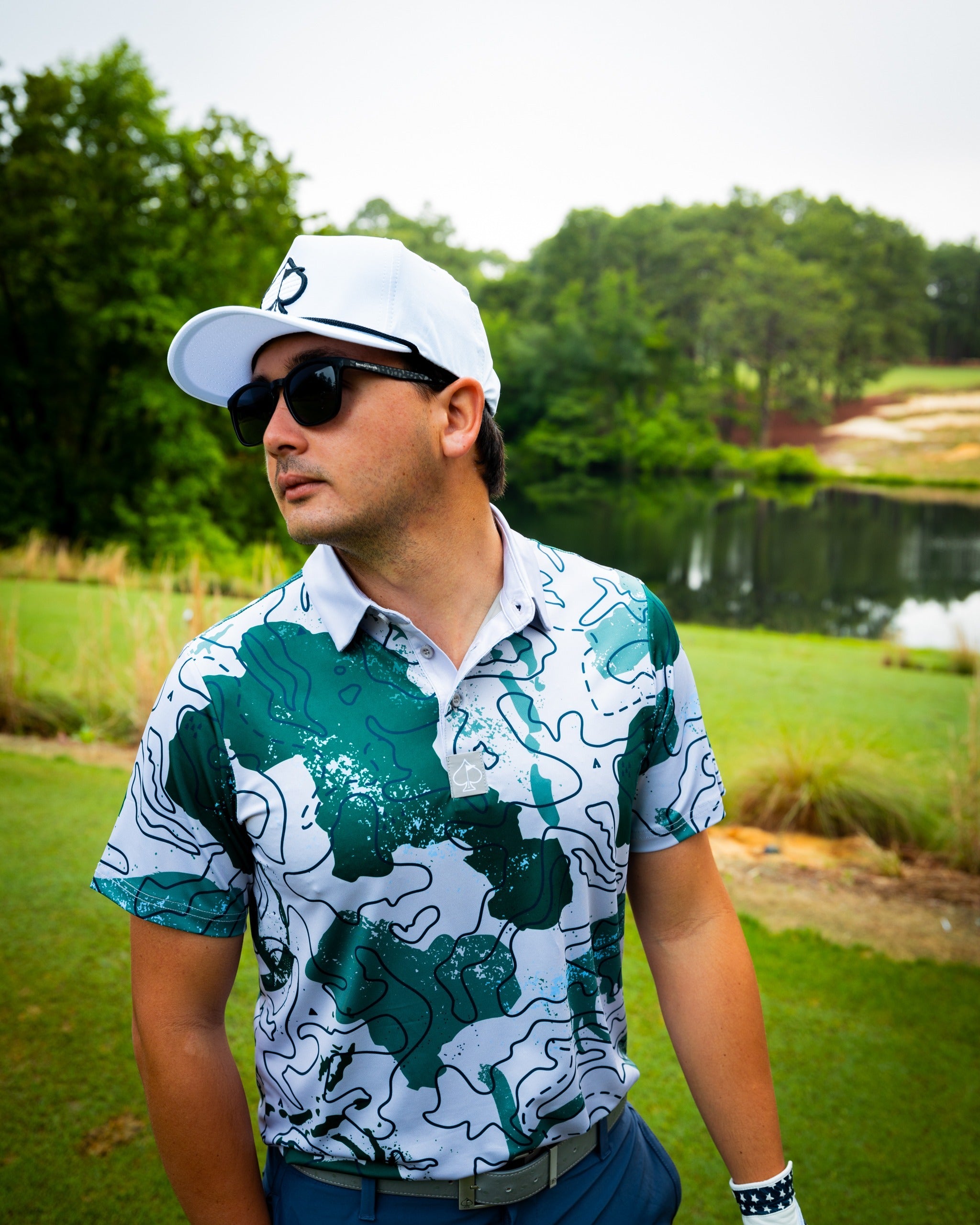 A golfer wearing the Yellowstone Topo polo shirt with a green and white topo map design, standing by a serene water feature.