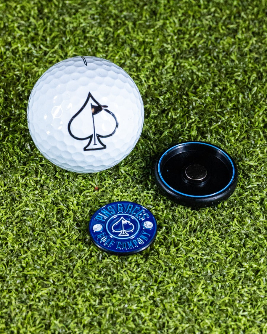 Navy pop-out medallion ball marker beside a golf ball with an ace of spades logo on a green turf background.