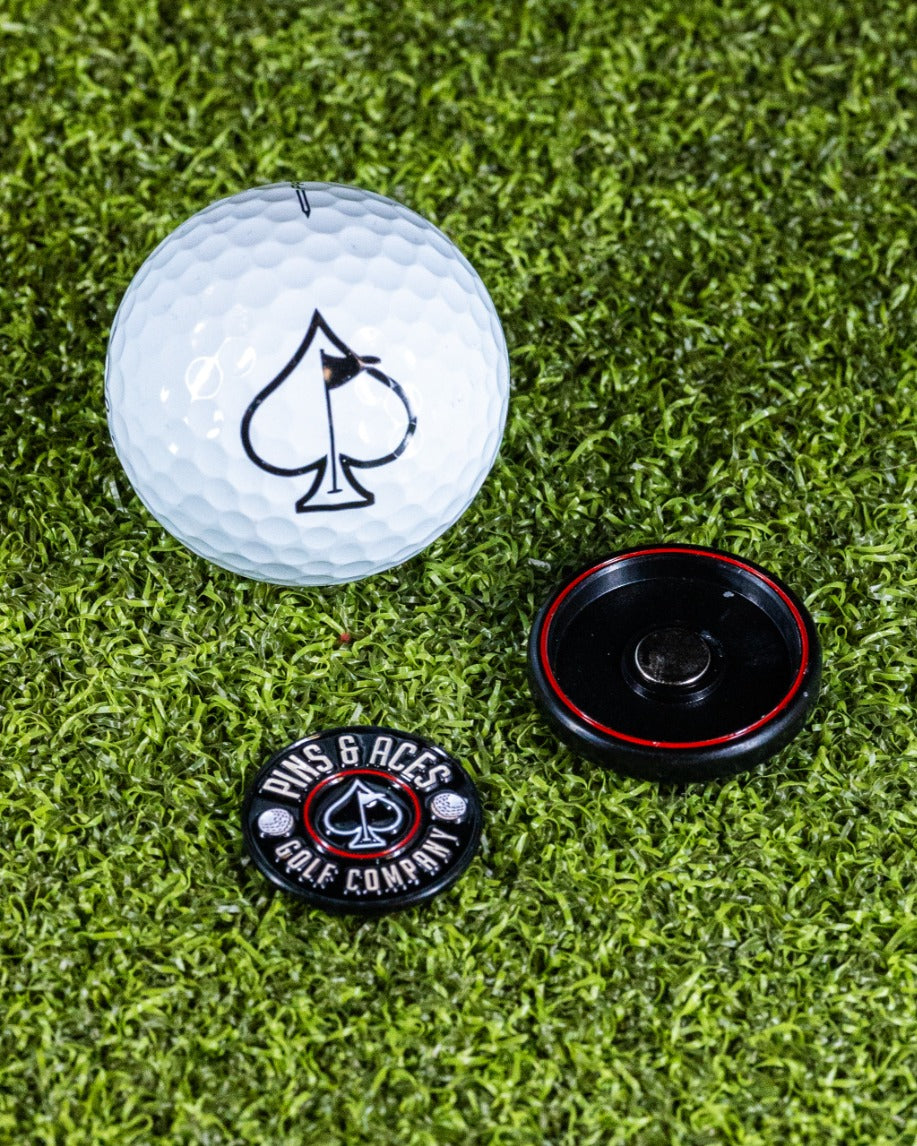 Black Pop Out Medallion Ball Marker next to a golf ball featuring a spade logo, set on green turf.