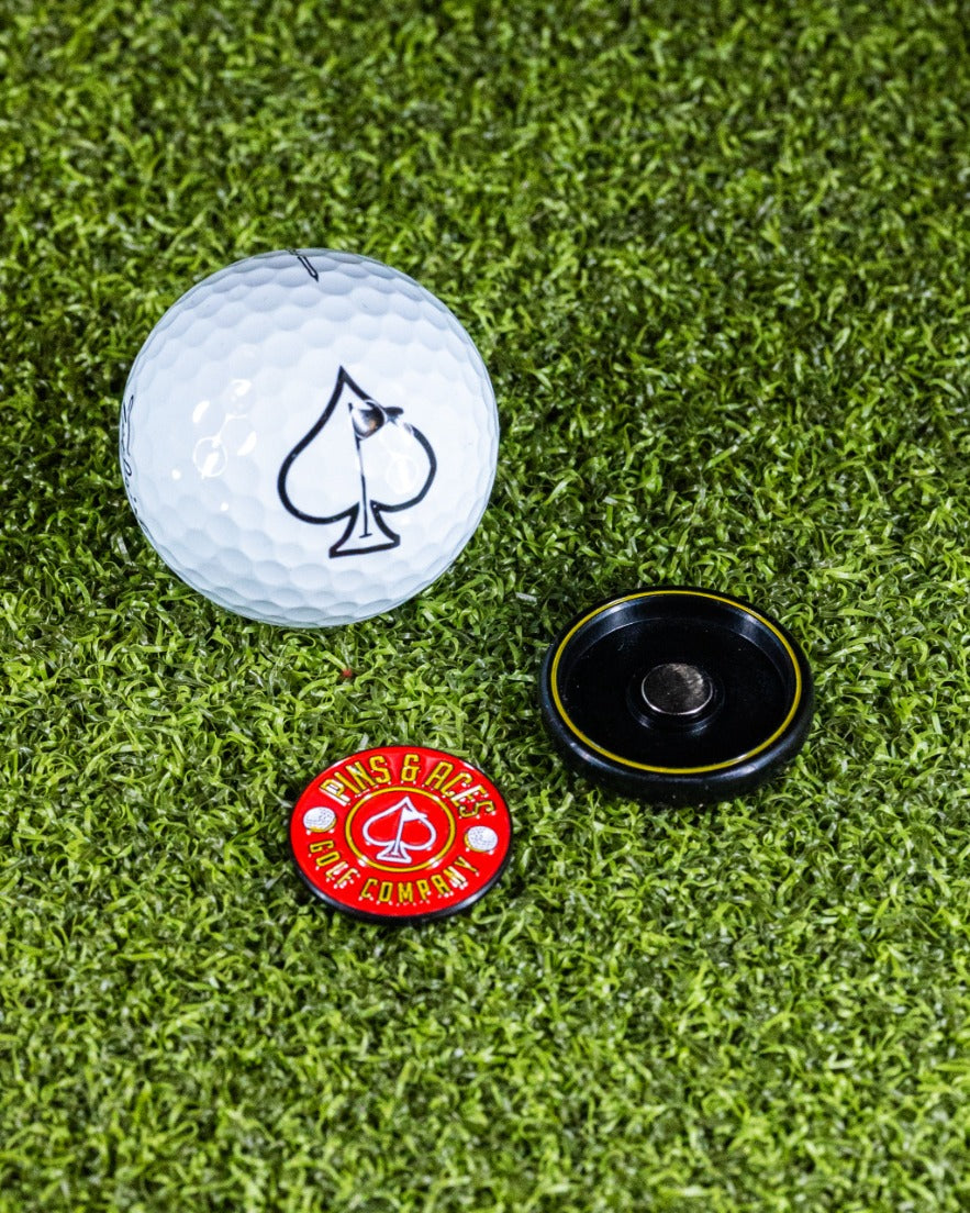 Red Pop Out Medallion Ball Marker next to a golf ball with an ace logo, set on green turf. Ideal for stylish golfers.