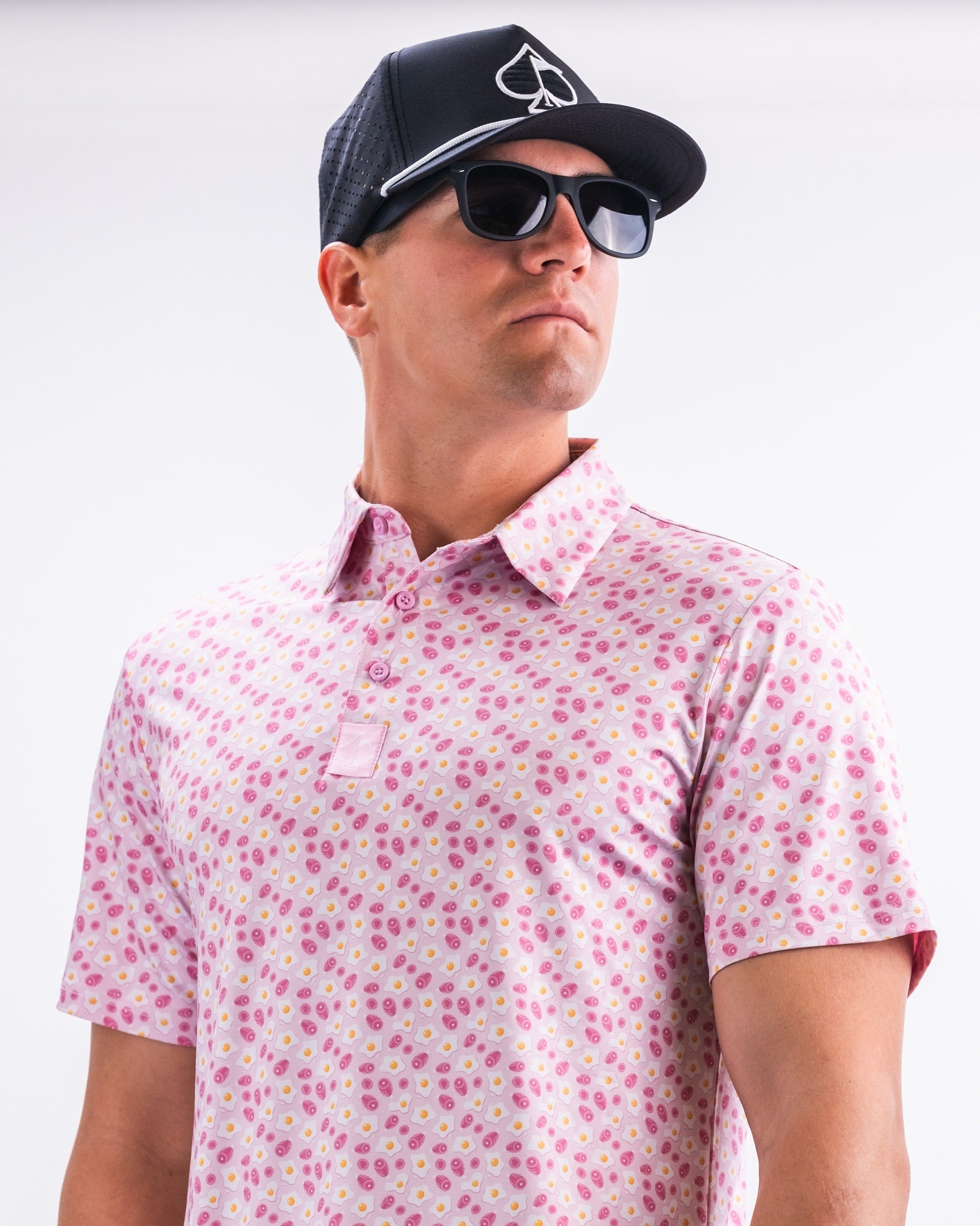 Stylish golfer wearing a pink patterned polo shirt with a modern fit, sunglasses, and a black cap, showcasing bold golf fashion.