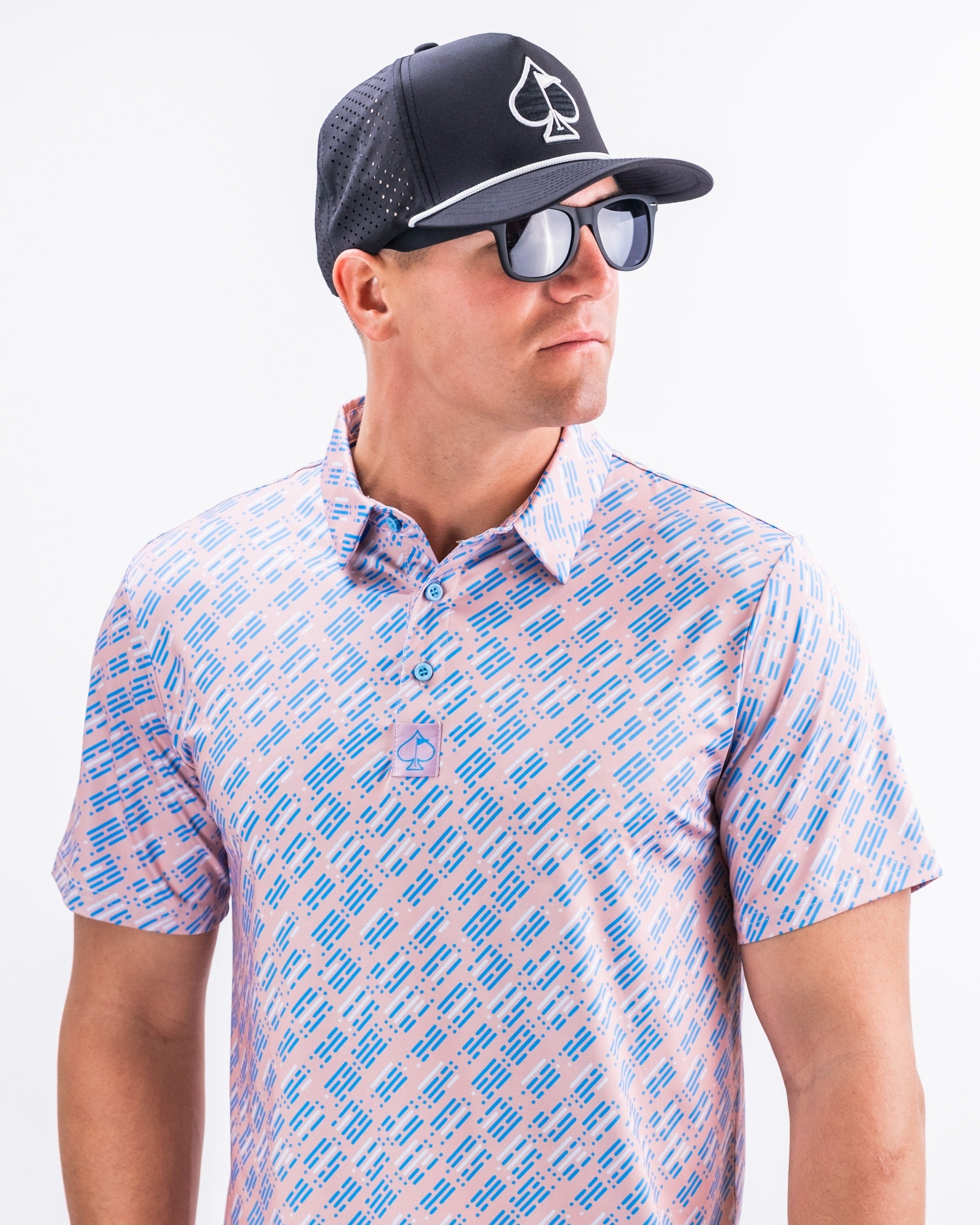 A golfer models the Skyline Dash Pink polo shirt, featuring a vibrant pattern, paired with a black cap and sunglasses.