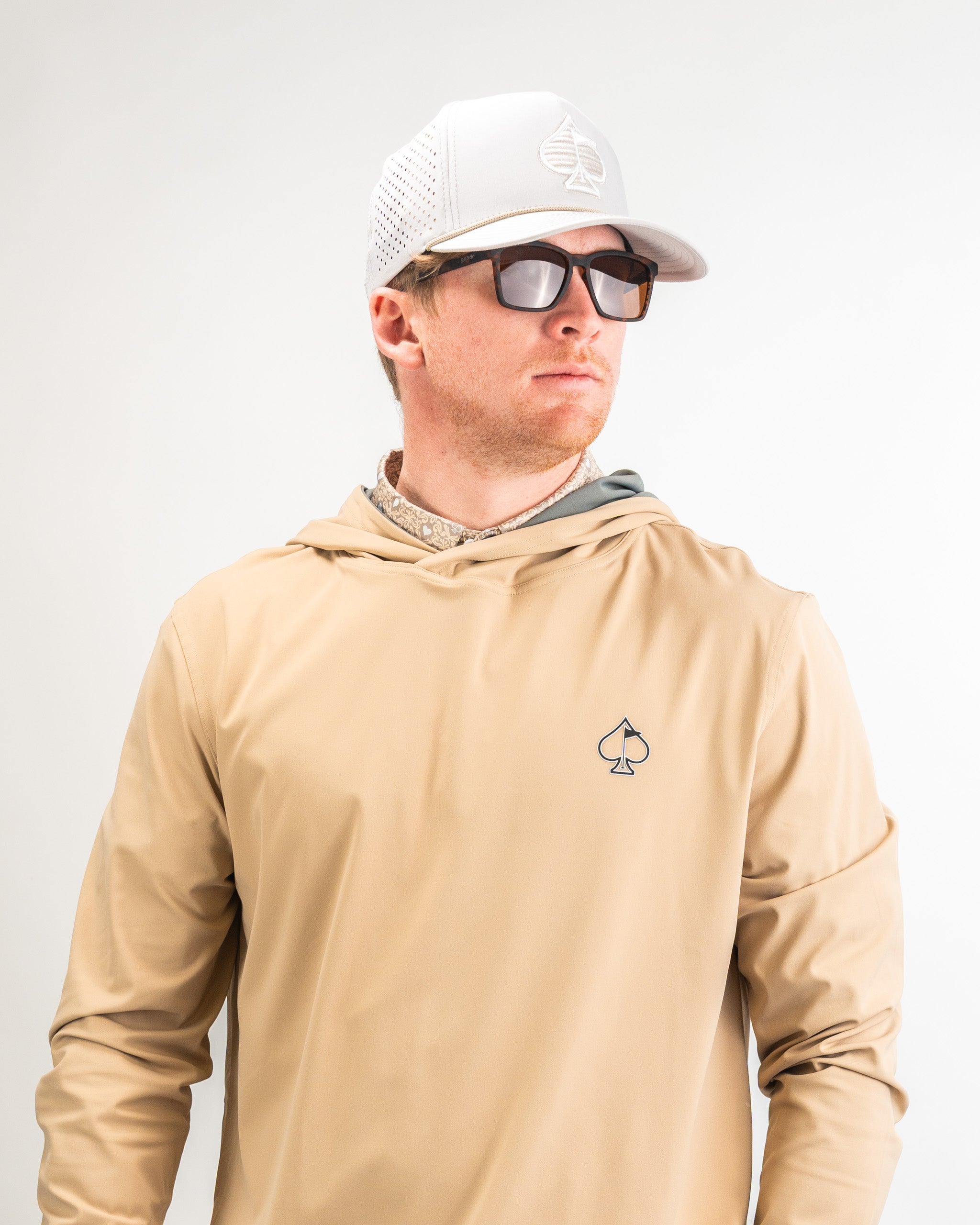 Performance Golf Hoodie in sand color, featuring a logo on the chest, paired with a light cap and sunglasses.