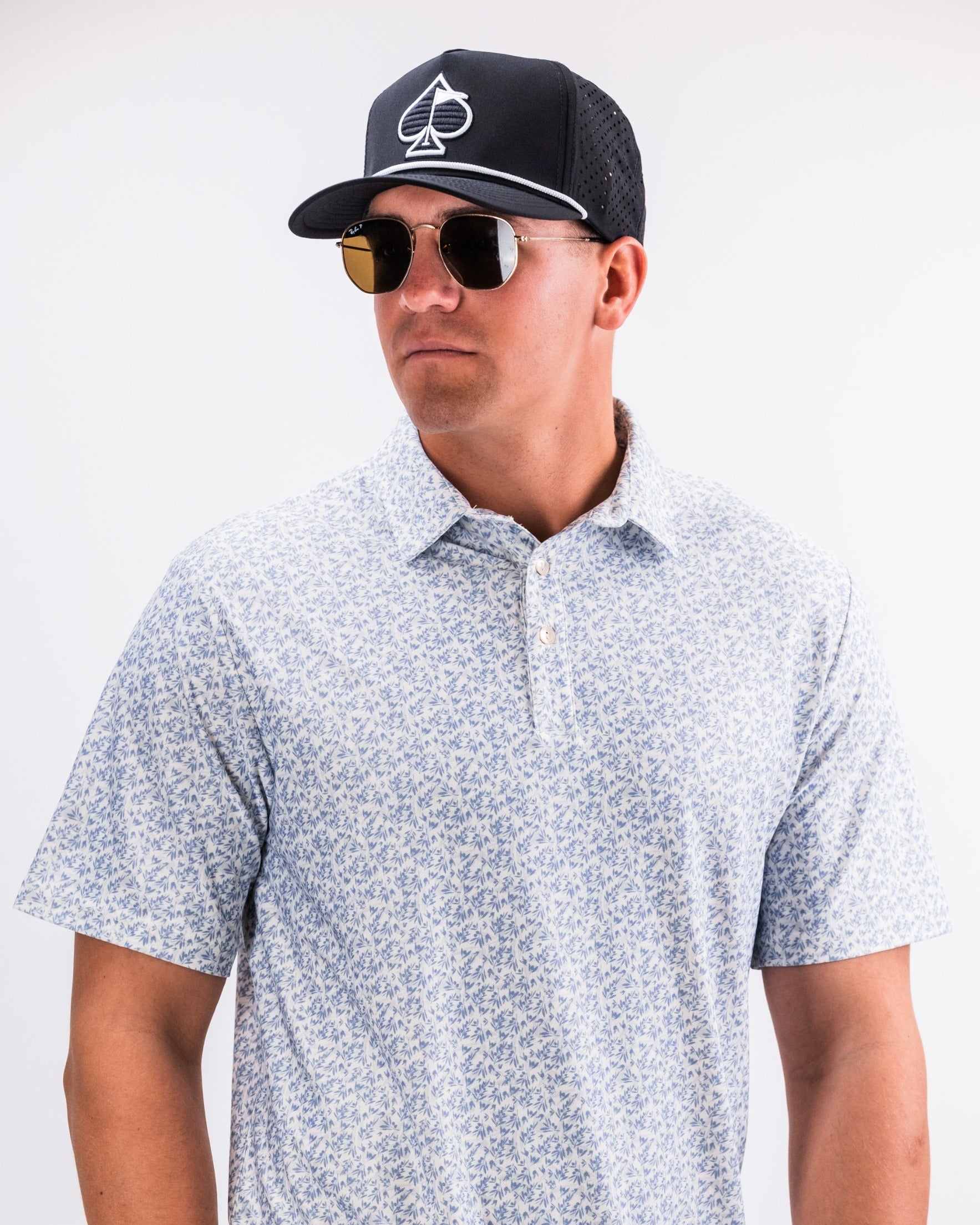 Stylish man wearing an olive green floral polo shirt with a black cap and sunglasses, showcasing bold golf fashion.