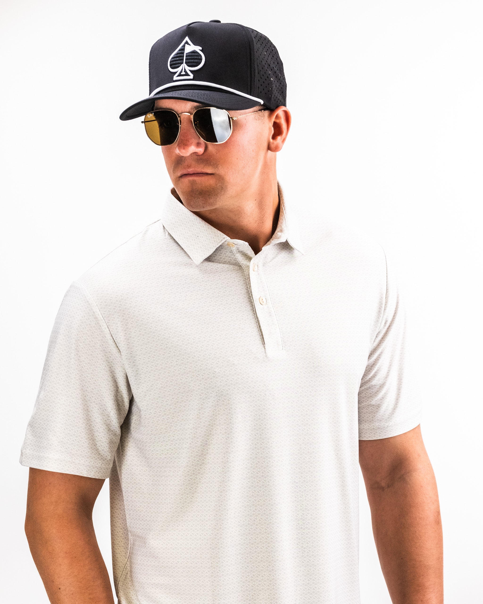 Stylish golfer wearing a light-colored Player Preferred Polo and a black cap with a spade logo, paired with sunglasses.