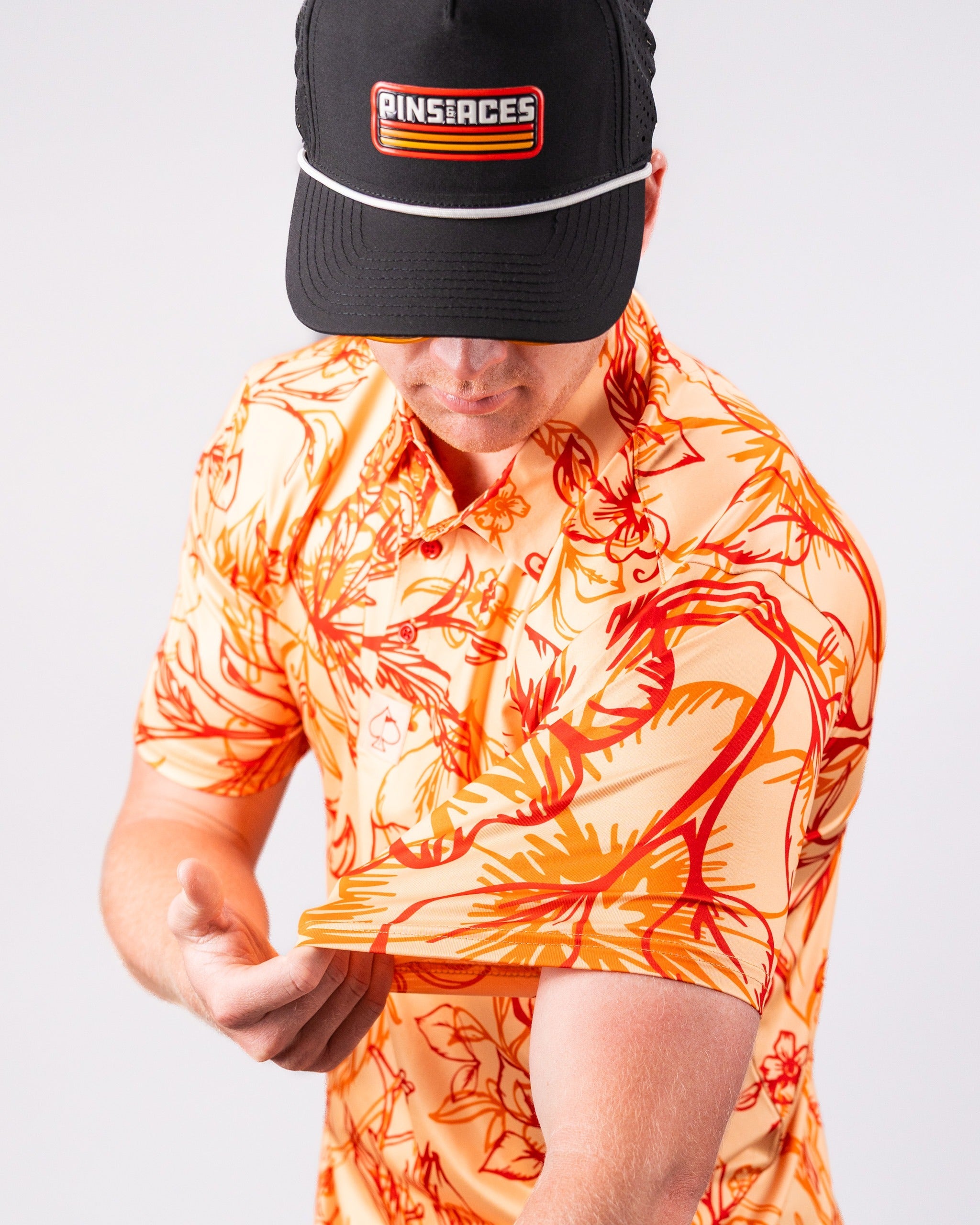 Bold orange and yellow floral golf shirt with a golfer adjusting the sleeve, wearing a black cap with a logo.