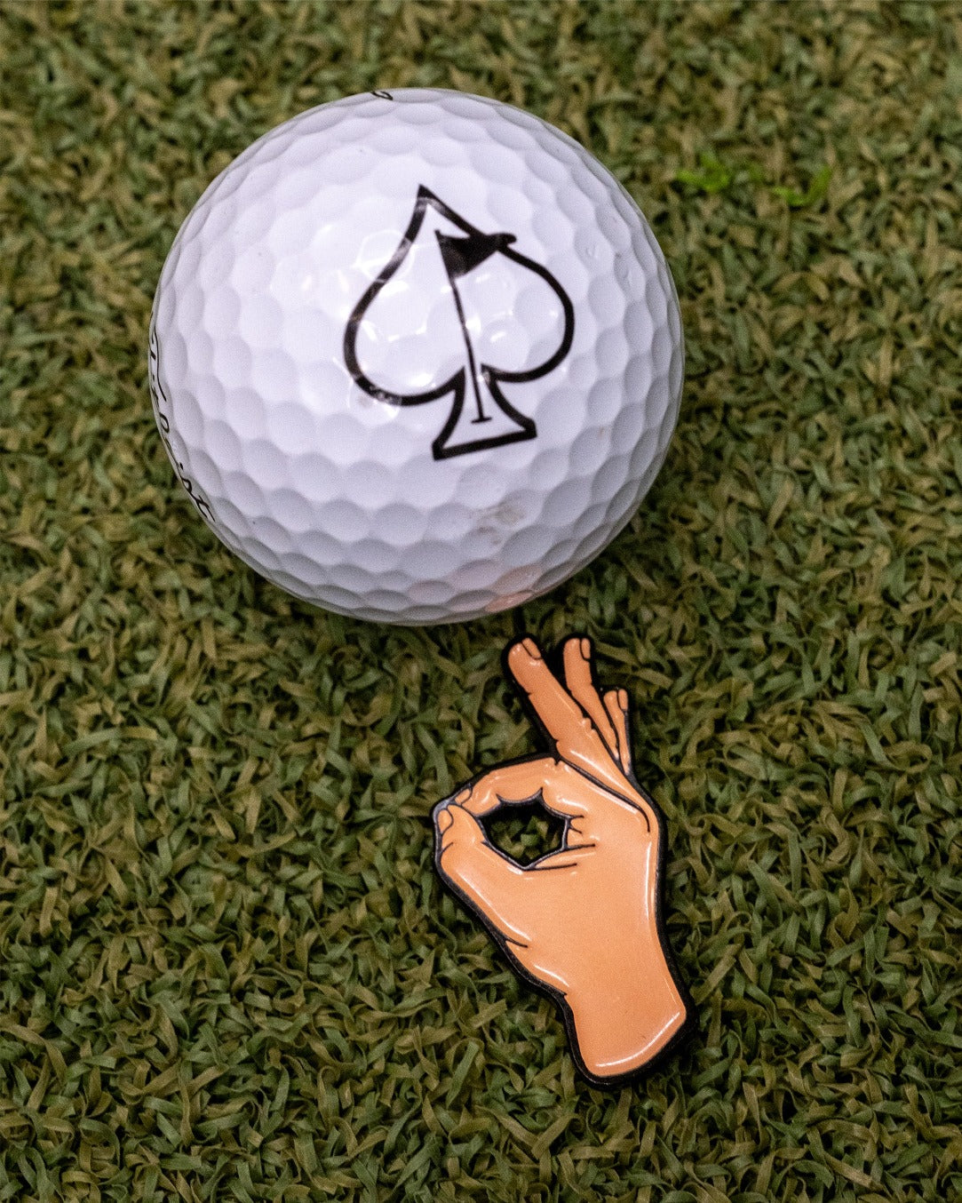 OK Hand Ball Marker next to a golf ball with an ace of spades logo, set against a green turf background.