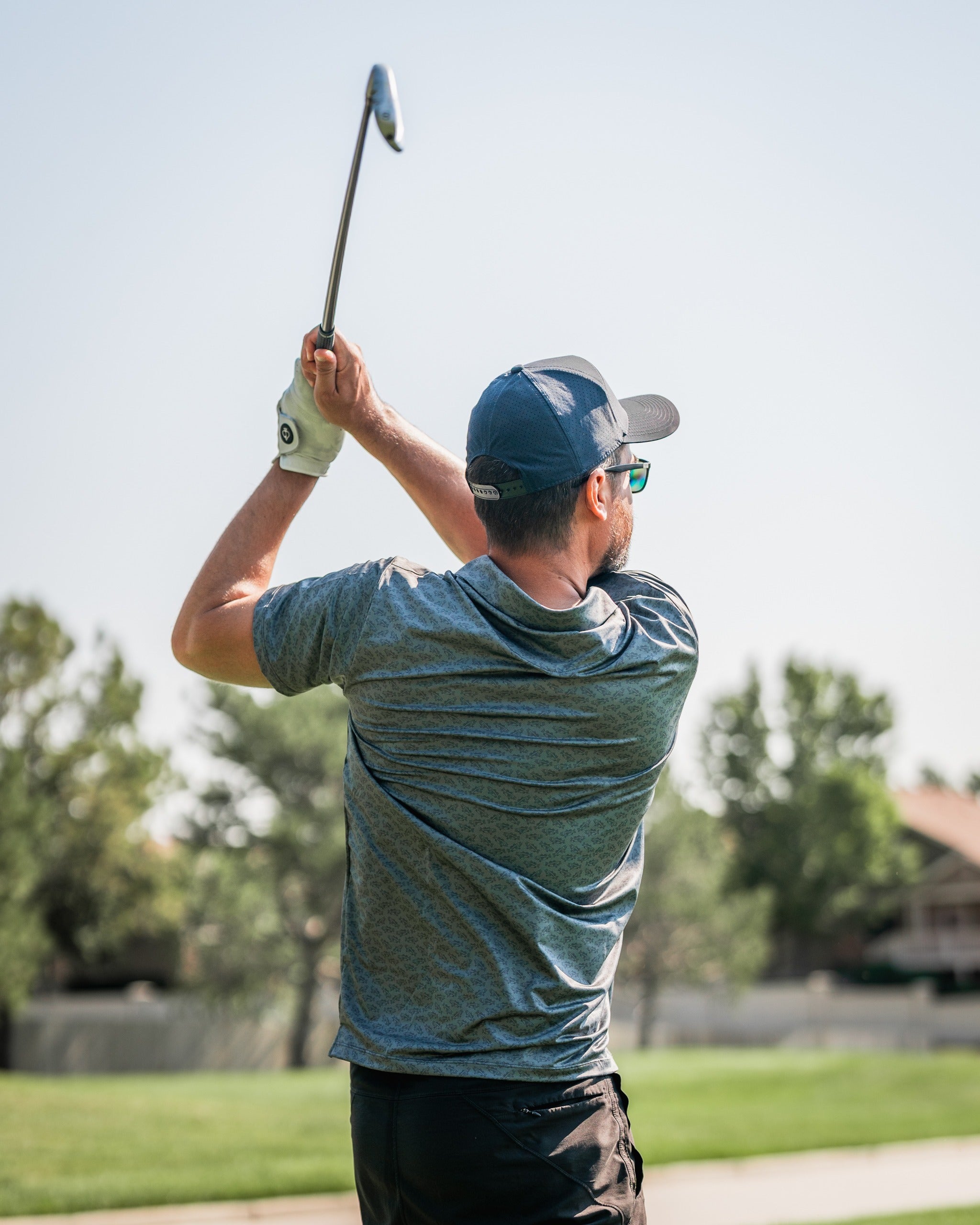 Golfer in a blue shirt and cap swings a club on a sunny course, showcasing the Large Mouth golf apparel.