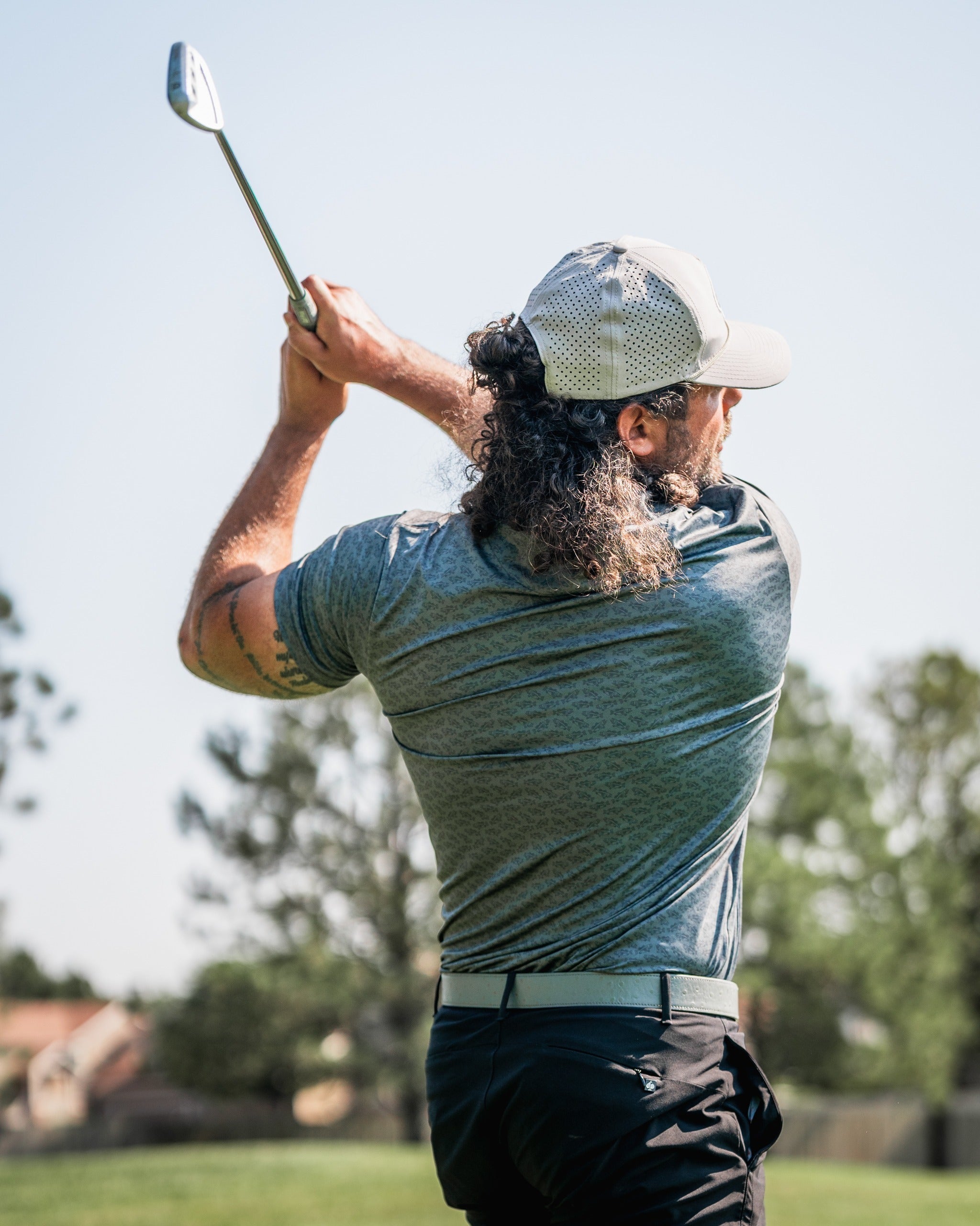 A golfer swings a club on the course, showcasing a stylish gray golf shirt and a cap, emphasizing bold athletic apparel.