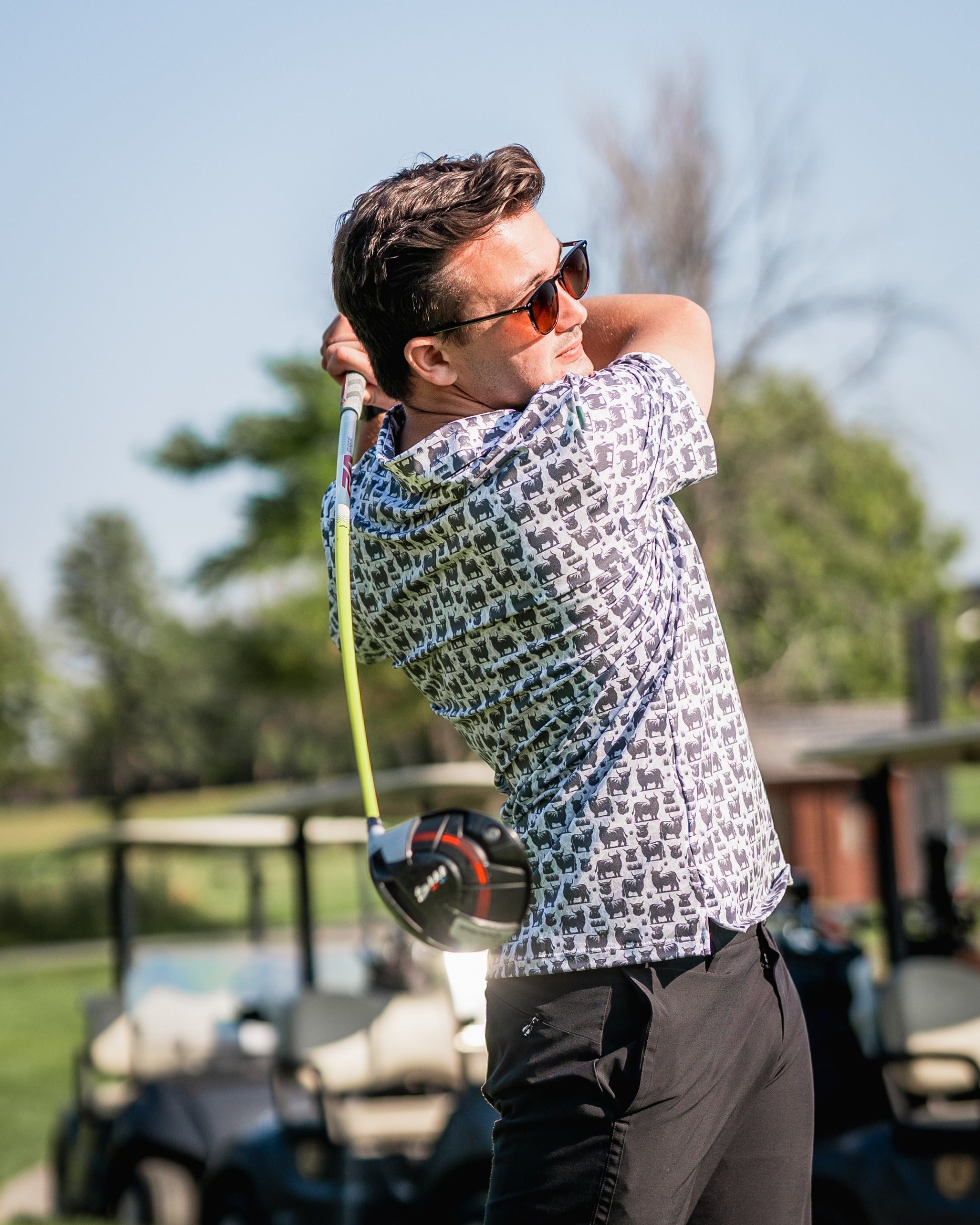 A golfer swings a driver while wearing a Highland Cow patterned shirt on a sunny course, showcasing bold golf fashion.