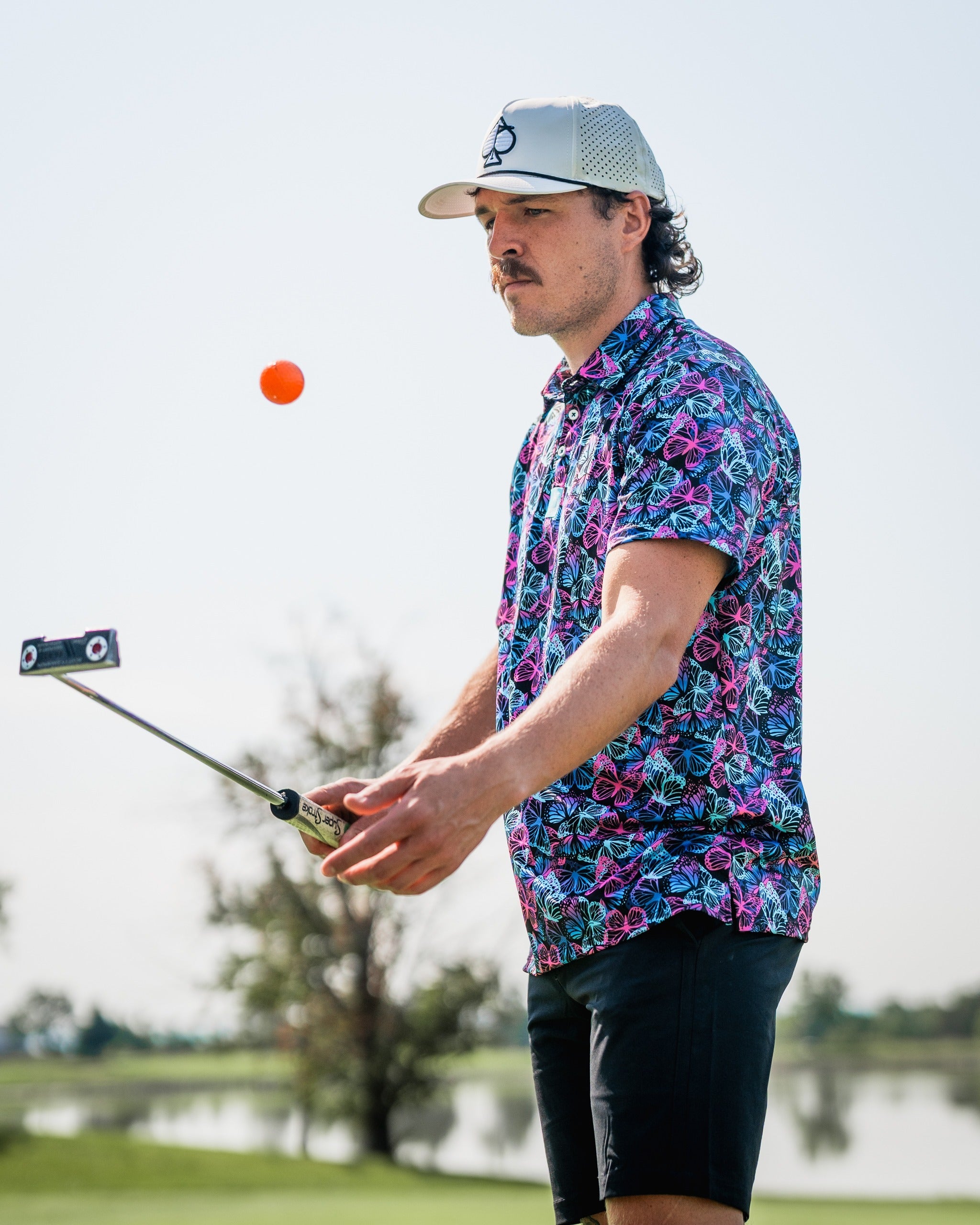 A golfer wearing a vibrant butterfly-patterned shirt prepares to putt on the course, with a bright orange ball in focus.