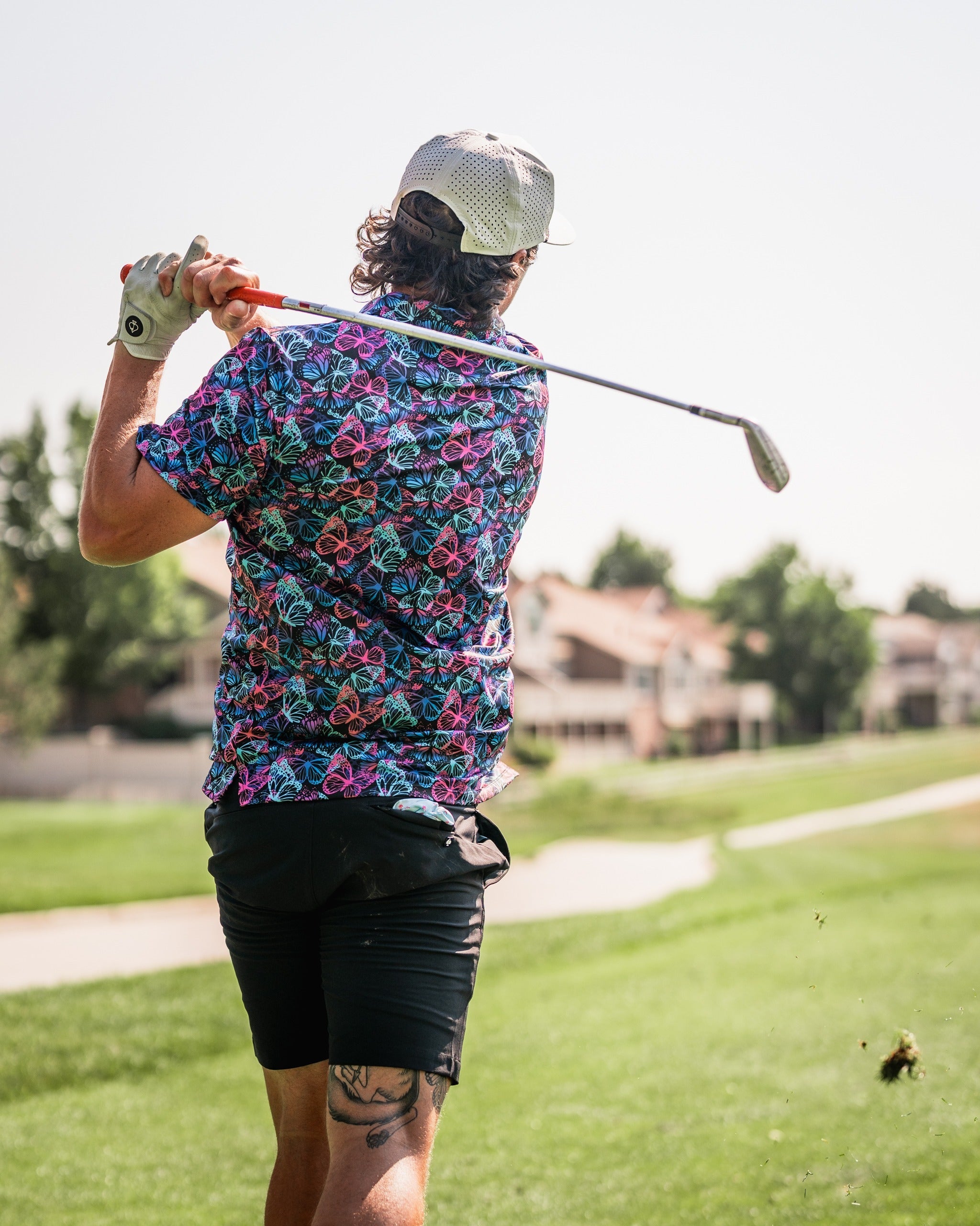 A golfer swings a club while wearing a vibrant butterfly-patterned shirt on a sunny course.