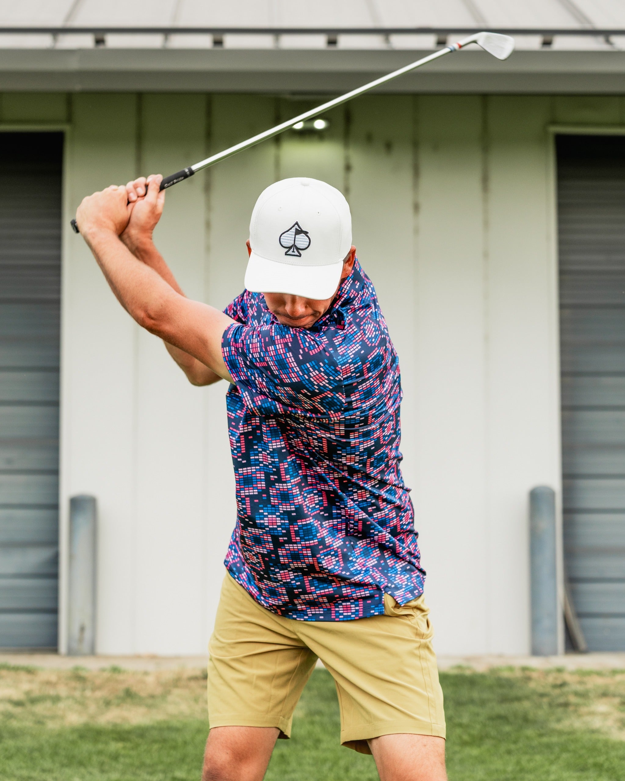 A golfer swings a club while wearing a vibrant Tetrafloral Dark shirt and beige shorts on a golf course.