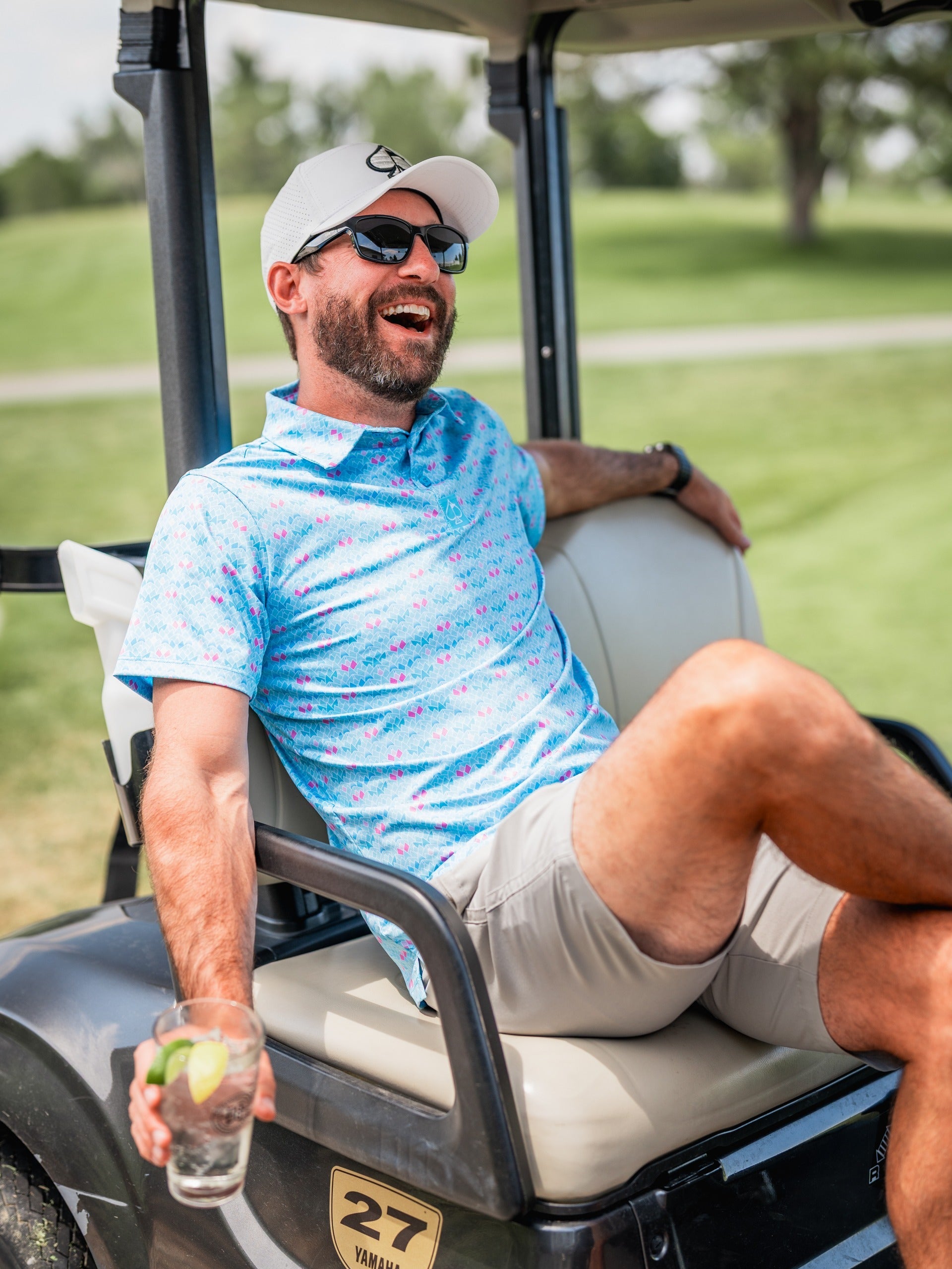 Man in a floral golf polo shirt relaxes on a golf cart, enjoying a drink on a sunny day at the golf course.