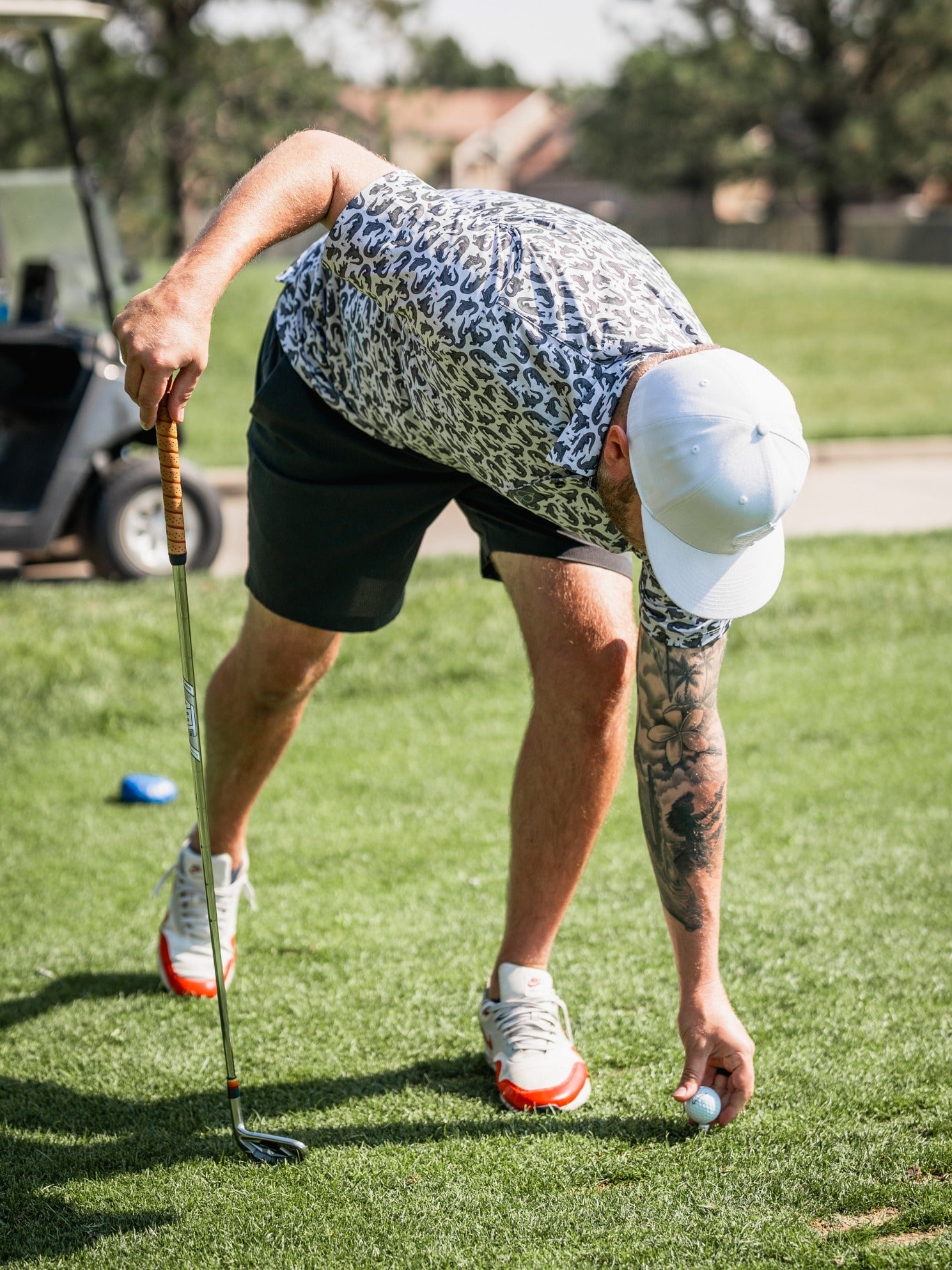 A golfer in a leopard print shirt and shorts bends down to pick up a golf ball on the course.