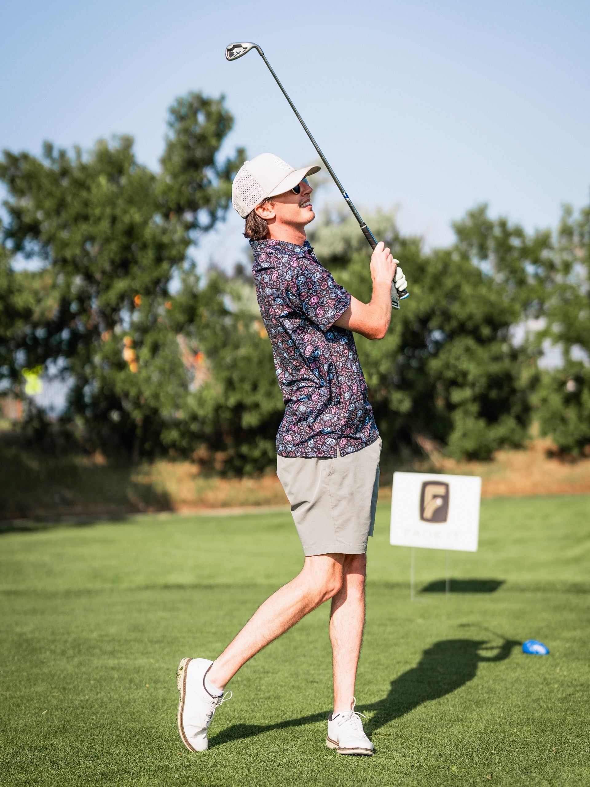 A golfer swings a club on the course, wearing a floral shirt and shorts, showcasing a stylish and relaxed golf look.