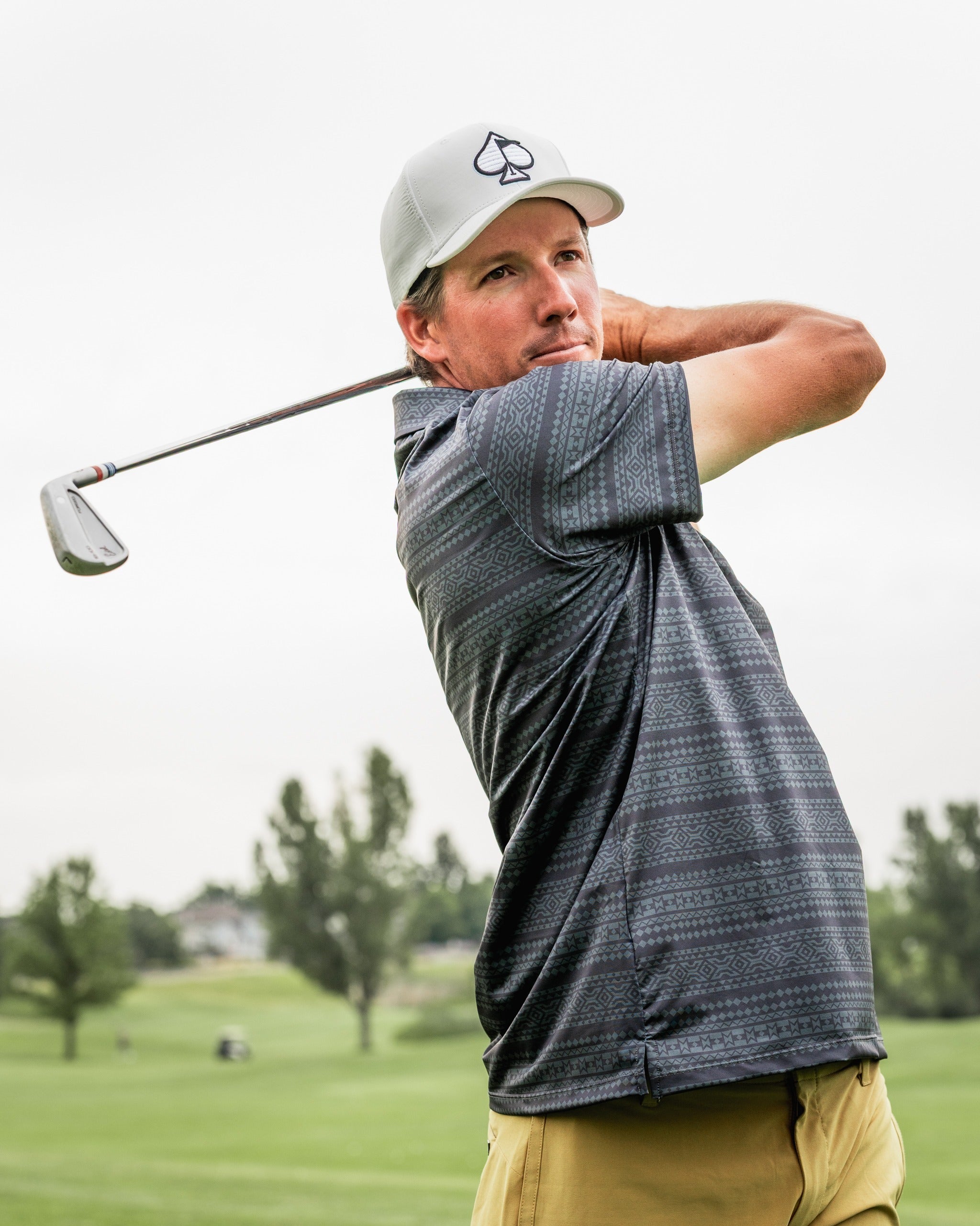 A golfer swings a club on the course, wearing a patterned polo shirt and a light gray cap, showcasing bold golf style.