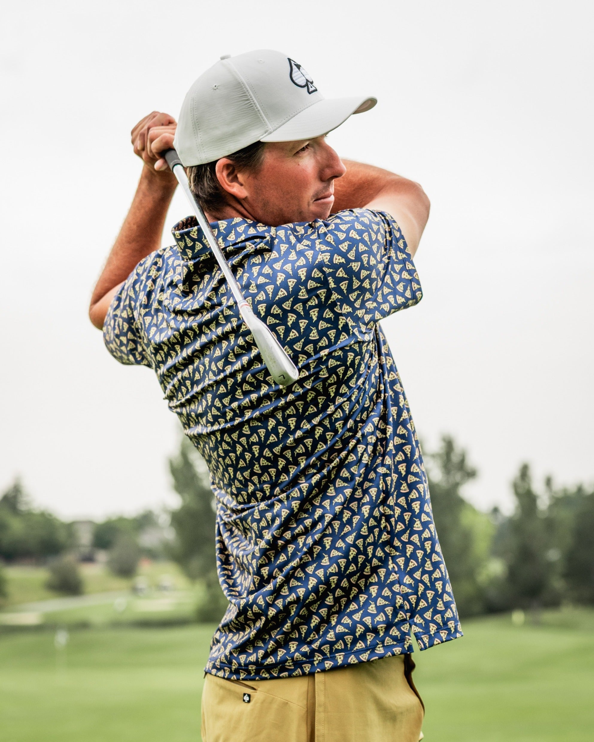 A golfer swings a club on the course, wearing a bold pizza-patterned shirt that showcases unique golf fashion.