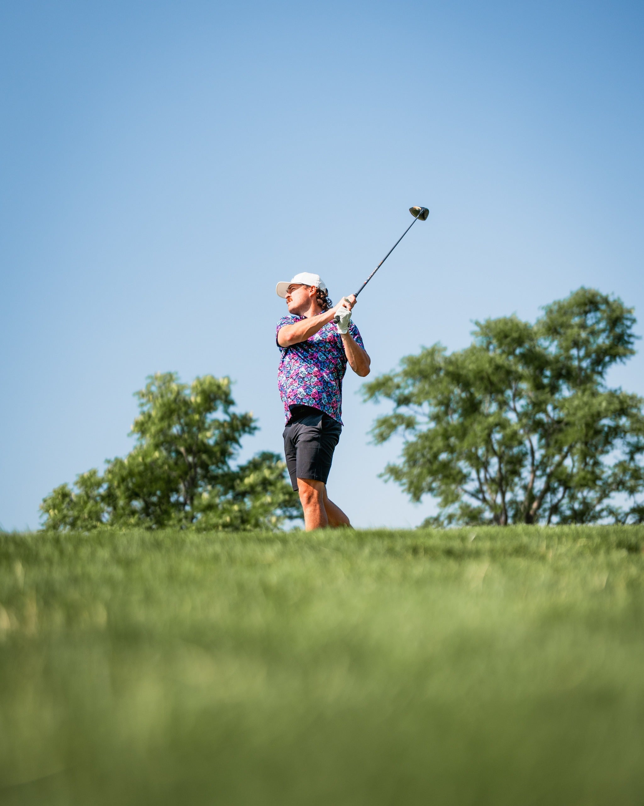 A golfer swings a driver on a sunny day, wearing a vibrant Electric Butterfly shirt and shorts, surrounded by greenery.