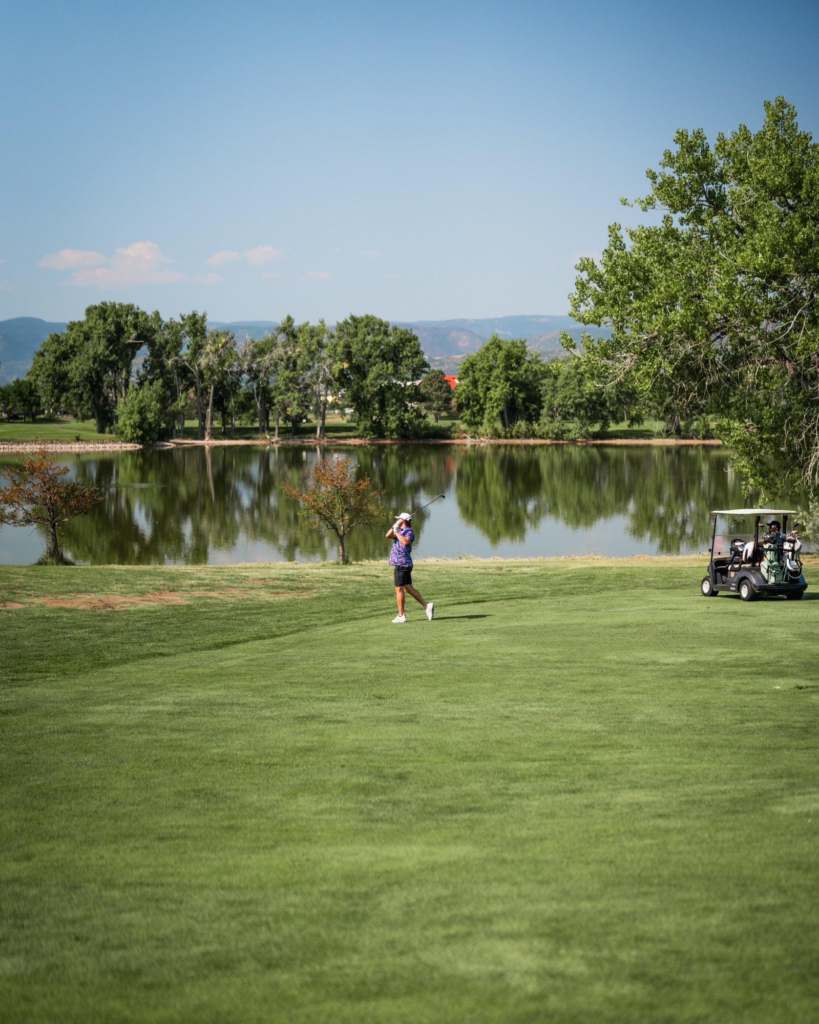 A golfer in a vibrant outfit walks on a lush green course near a serene lake, with a golf cart parked nearby.