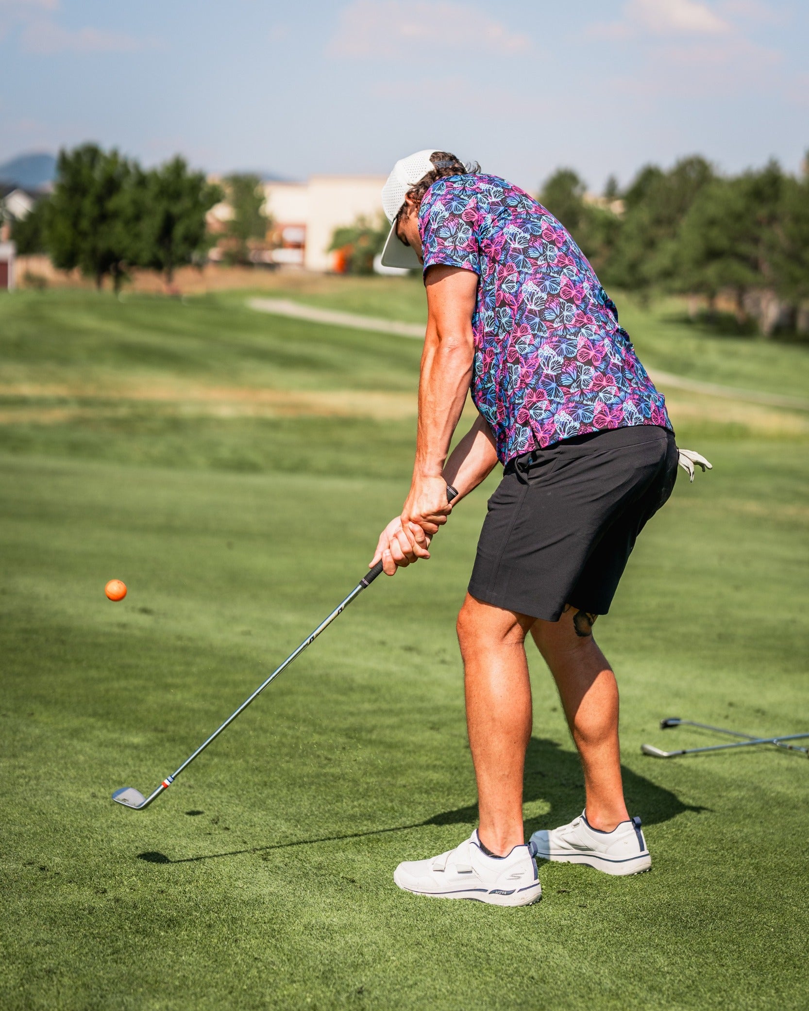 A golfer in a vibrant floral shirt and shorts swings a club on a sunny course, preparing to hit a golf ball.