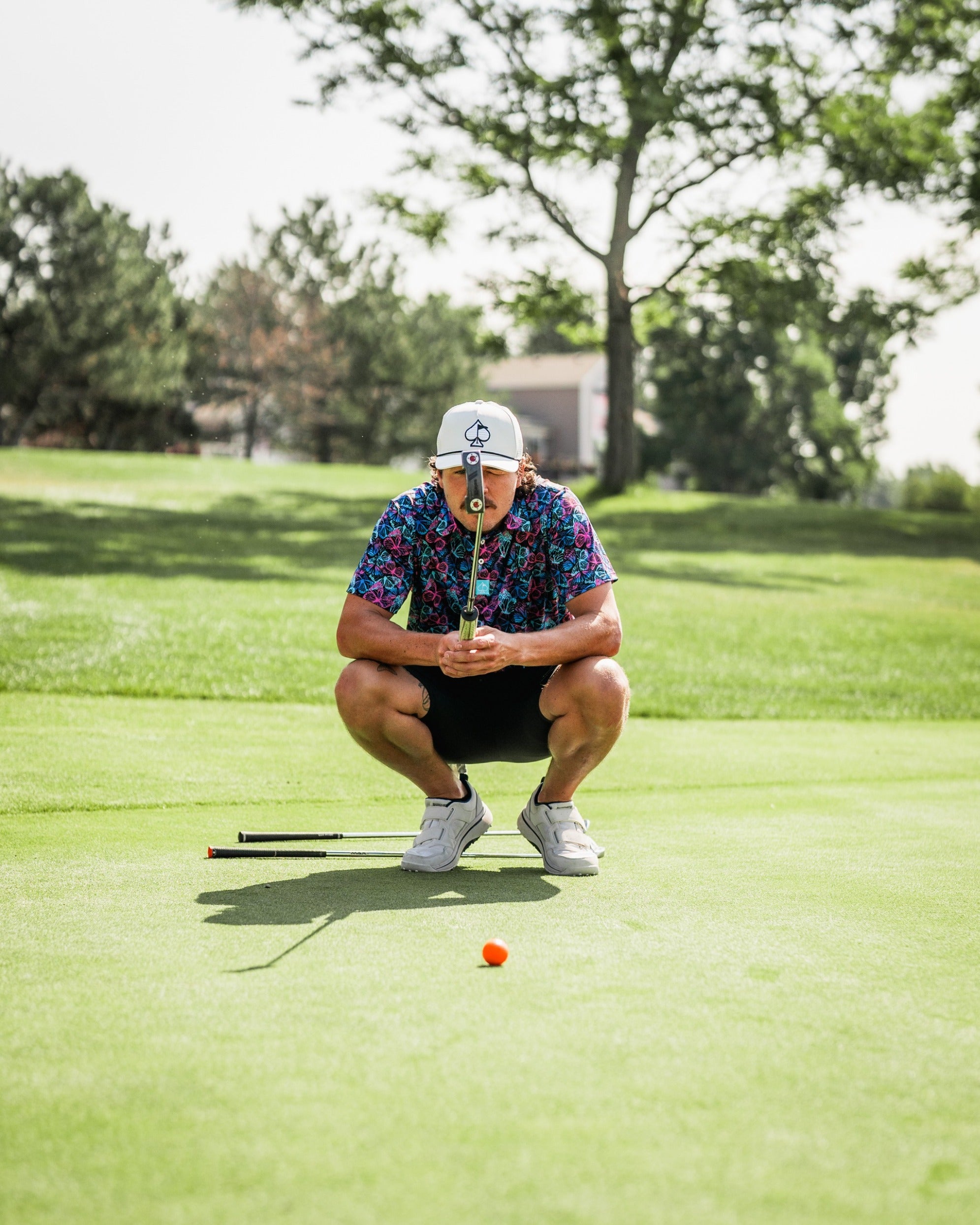 A golfer in a colorful floral shirt crouches on the green, focusing intently on a putt with an orange ball.