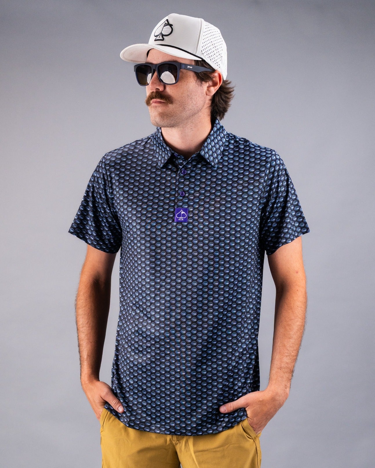Man wearing a navy patterned polo shirt with a button-up collar, sunglasses, and a white cap, posing confidently.