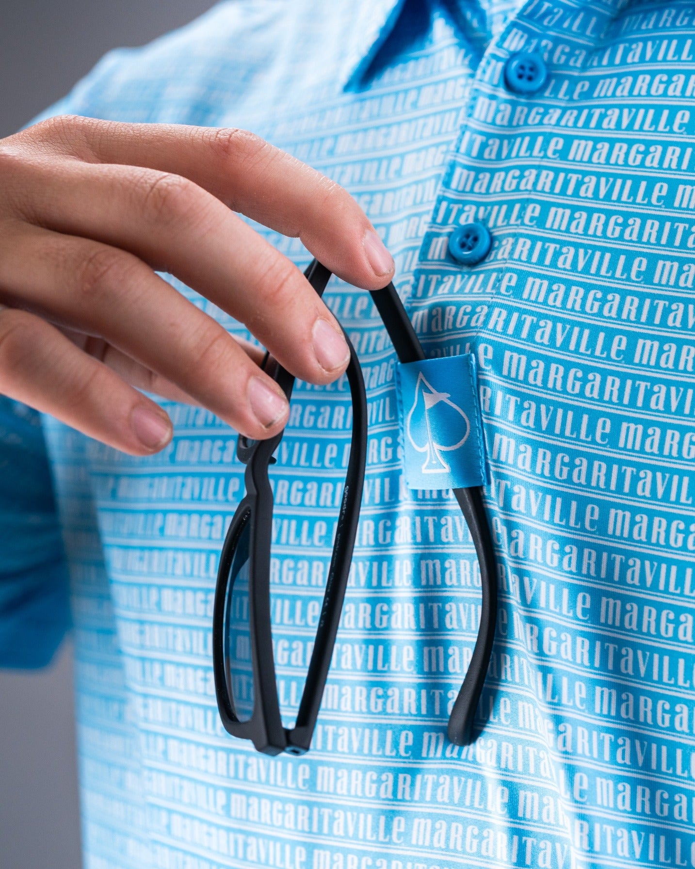 Light blue Margaritaville polo shirt featuring a repeating logo pattern, with sunglasses hanging from the collar.