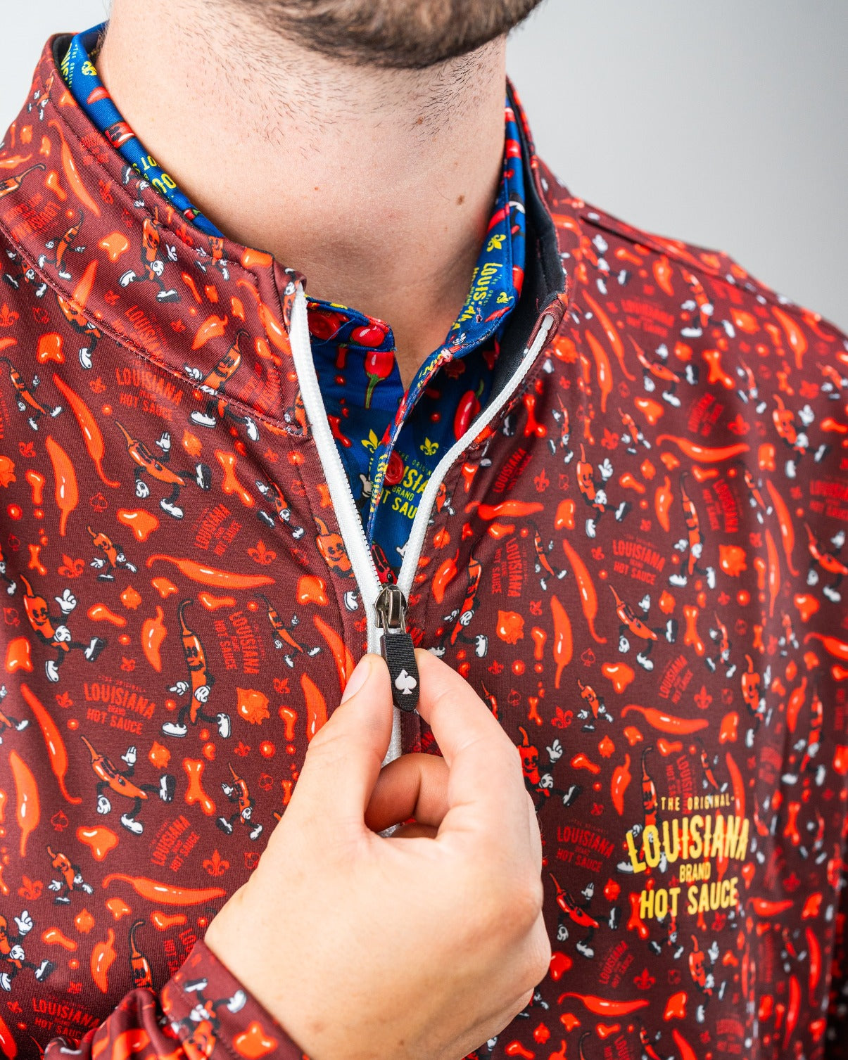 Close-up of a man wearing a red, chili pepper-patterned golf jacket with "Louisiana Hot Sauce" branding, partially unzippe...