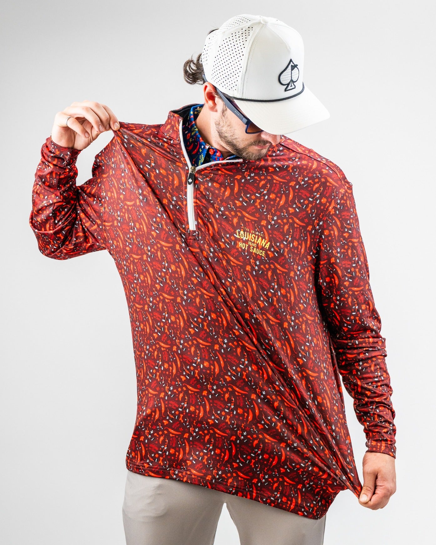 Man in a red, patterned golf jacket with "Louisiana Hot Sauce" logo, white cap with spade symbol, and sunglasses, showcasi...