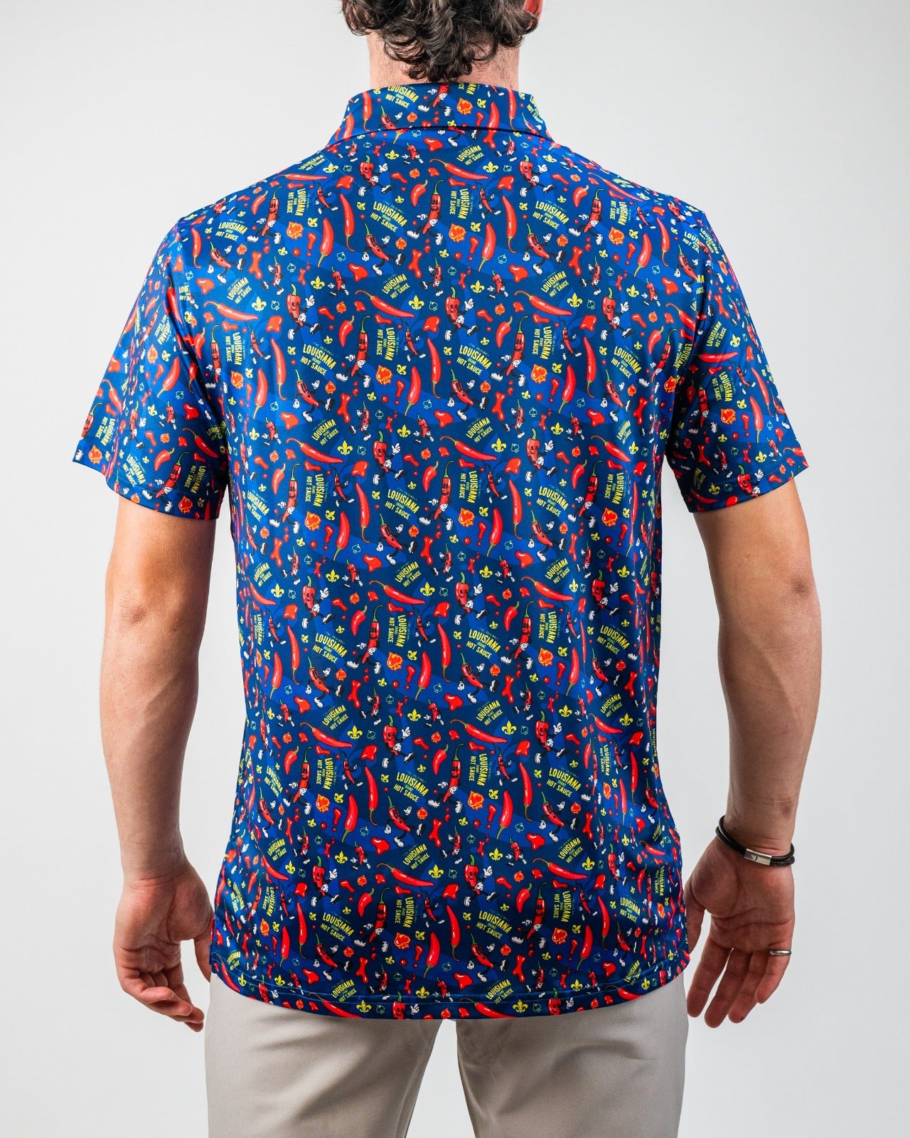 Back view of a man wearing a vibrant, blue, Louisiana-themed golf shirt with red, yellow, and green chili pepper, hot sauc...