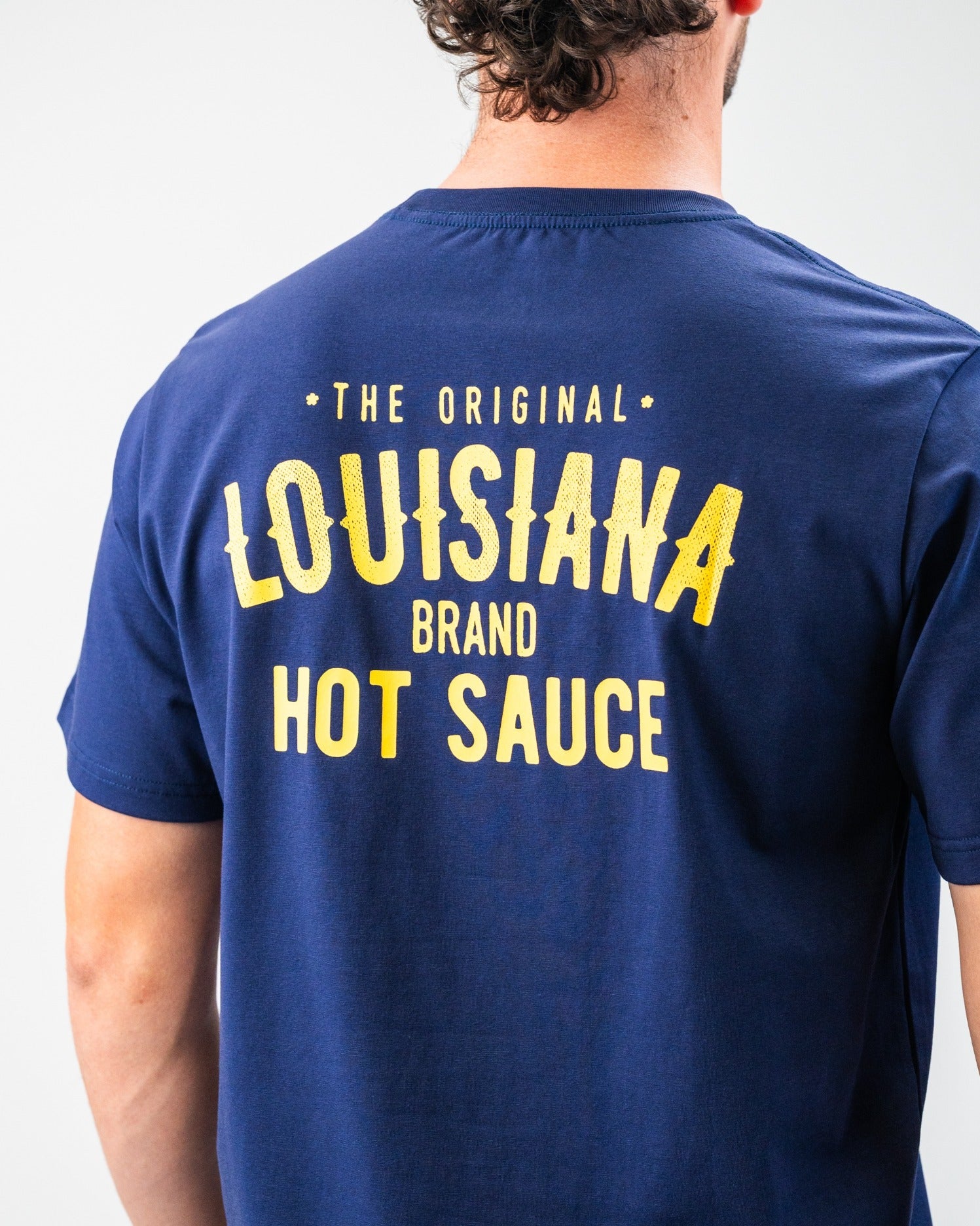 Back of navy golf t-shirt with bold yellow text: "The Original Louisiana Brand Hot Sauce" for standout golf style.