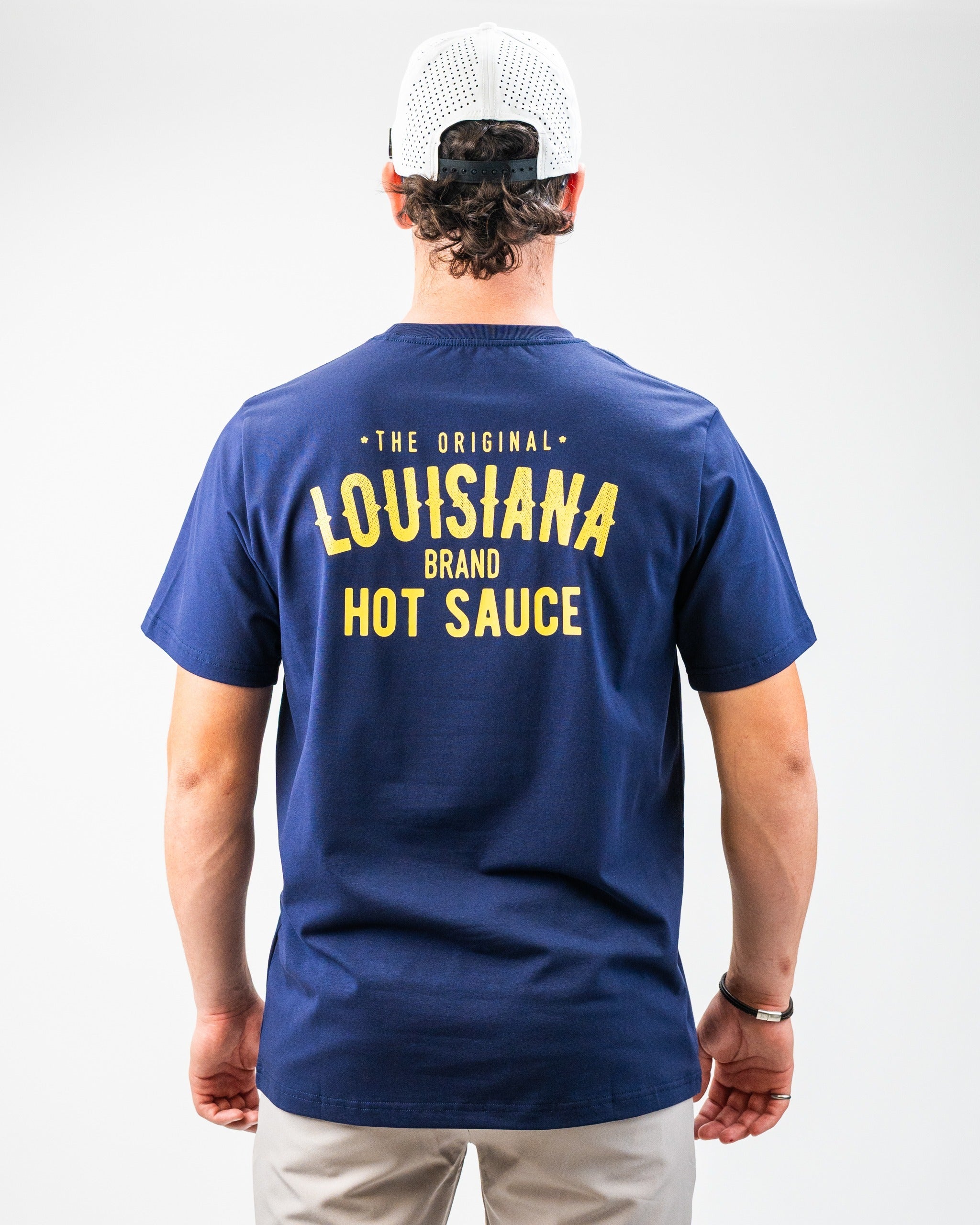 Back view of a golfer wearing a navy blue T-shirt with bold yellow "Louisiana Hot Sauce" text and a white mesh cap with bl...