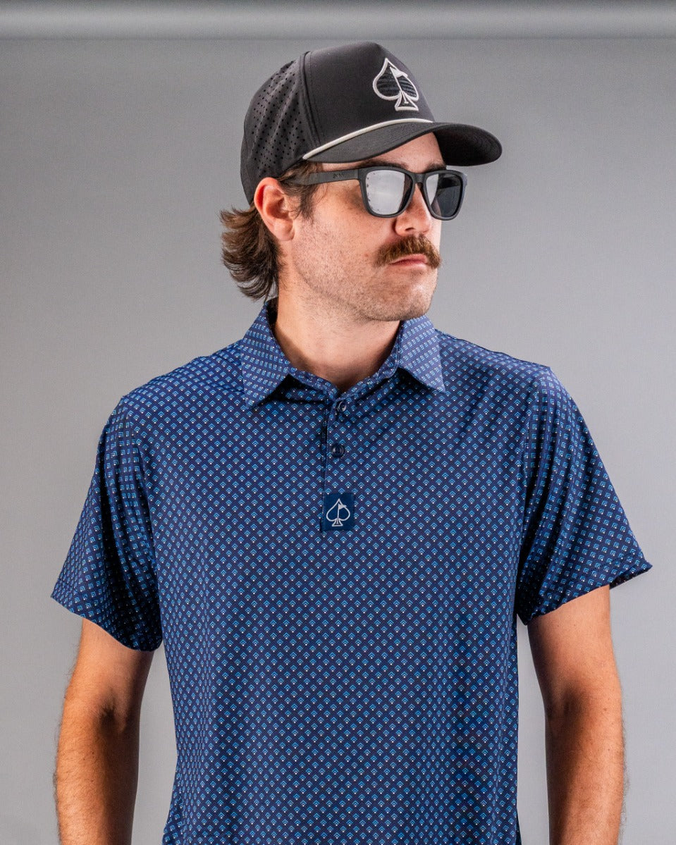 Man in blue patterned golf shirt, black cap with spade logo, and sunglasses, showcasing bold golf apparel for standout pla...