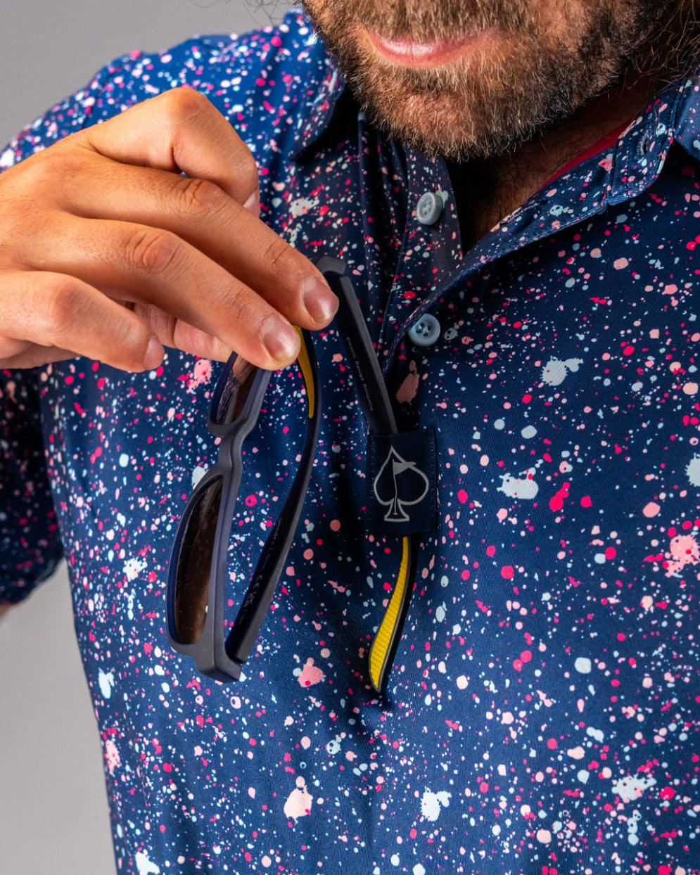 Man holding sunglasses while wearing a blue splatter-patterned golf shirt, showcasing bold and vibrant style.