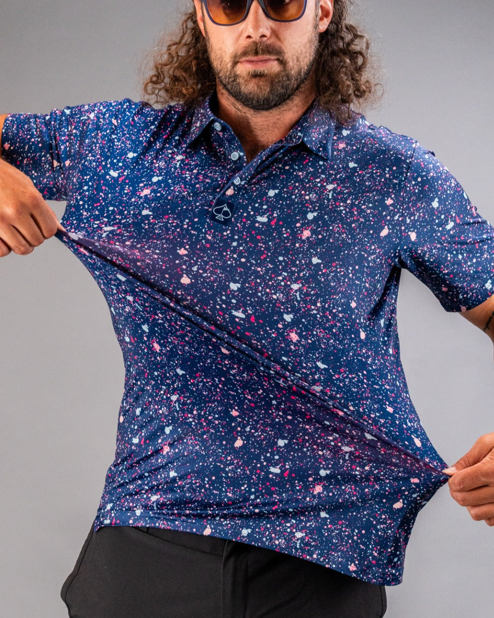 A golfer showcases a blue splatter polo shirt, featuring vibrant pink and white splashes against a navy background.