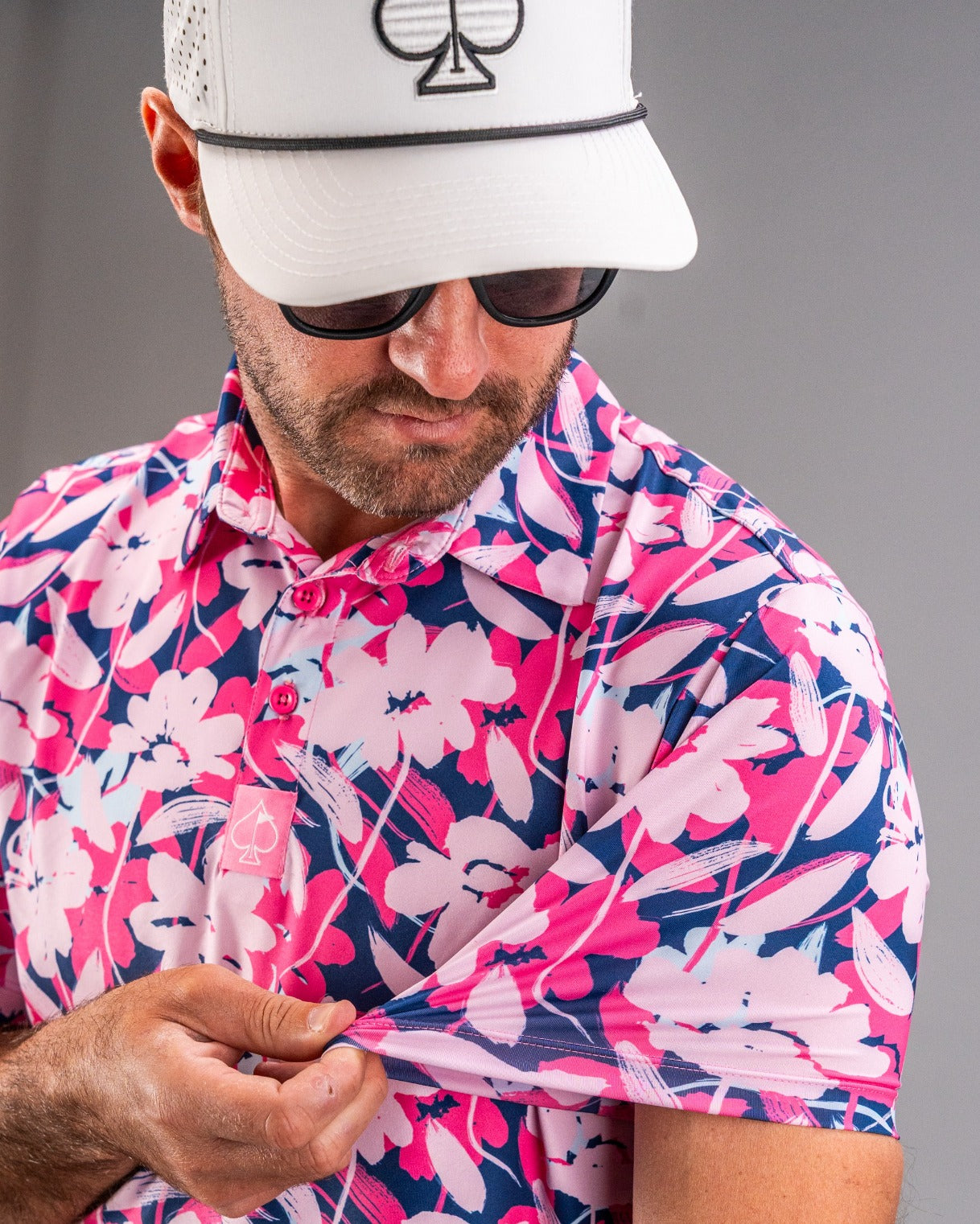 Man in pink, navy, and white floral golf shirt with black sunglasses and white cap featuring a spade symbol.