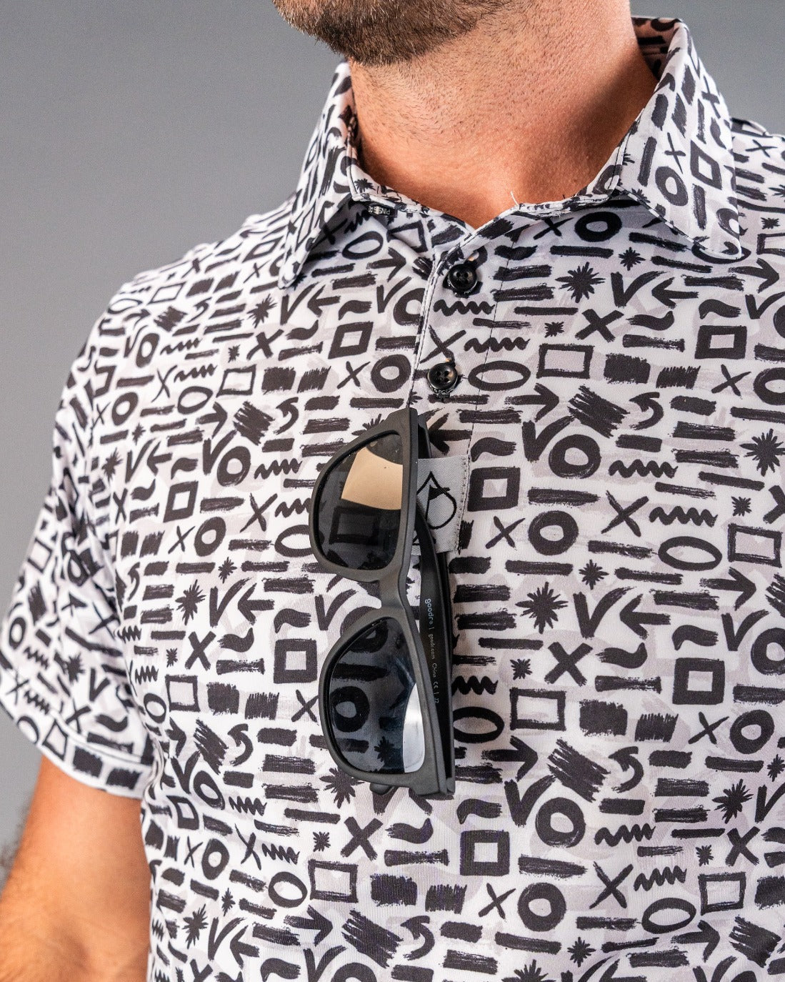 Black sunglasses hanging from a black-and-white patterned golf shirt with abstract symbols and bold design elements.